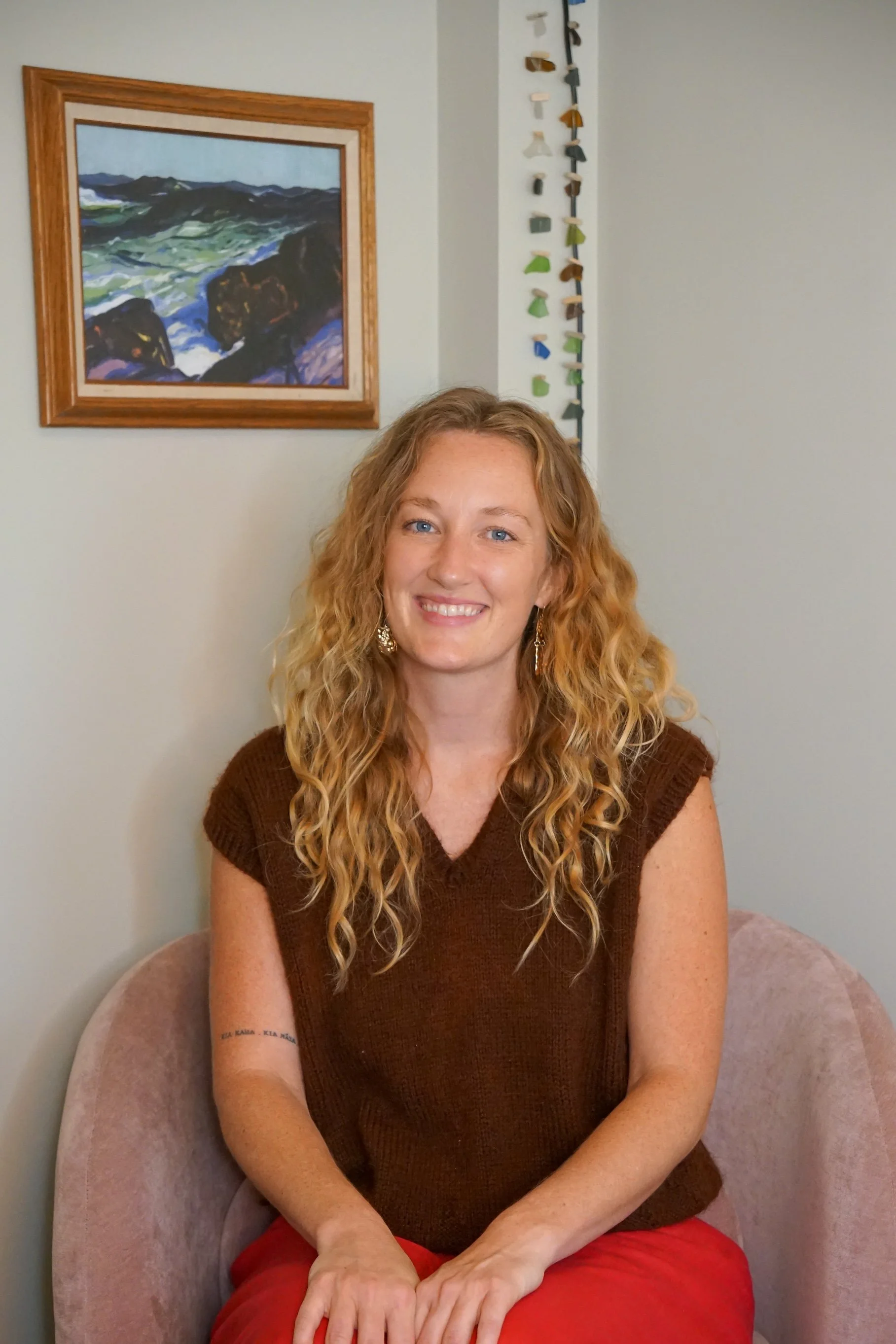 A woman with curly blonde hair, wearing earrings, a brown top, and red pants, sitting on a pink chair with a light green wall behind her. There is a framed landscape painting and a hanging decorative piece on the wall.