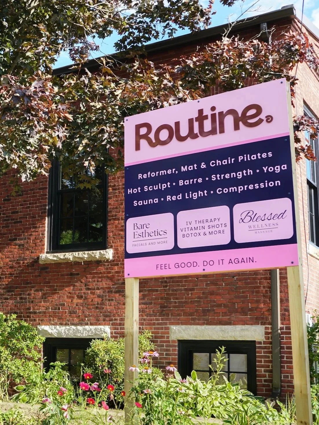 A large pink and navy sign for Routine Wellness Studio, situated in front of a brick building with windows and surrounded by flowers and trees.