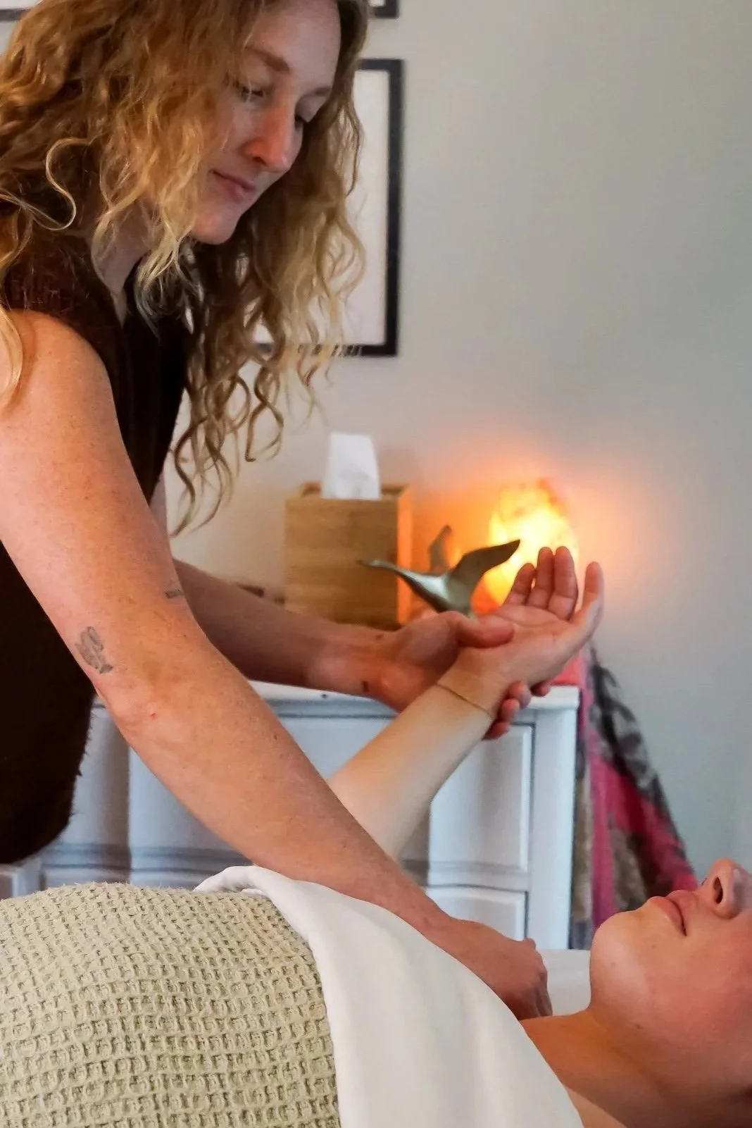 Woman giving massage to a person lying on a massage table in a room with soft lighting and salt lamp.