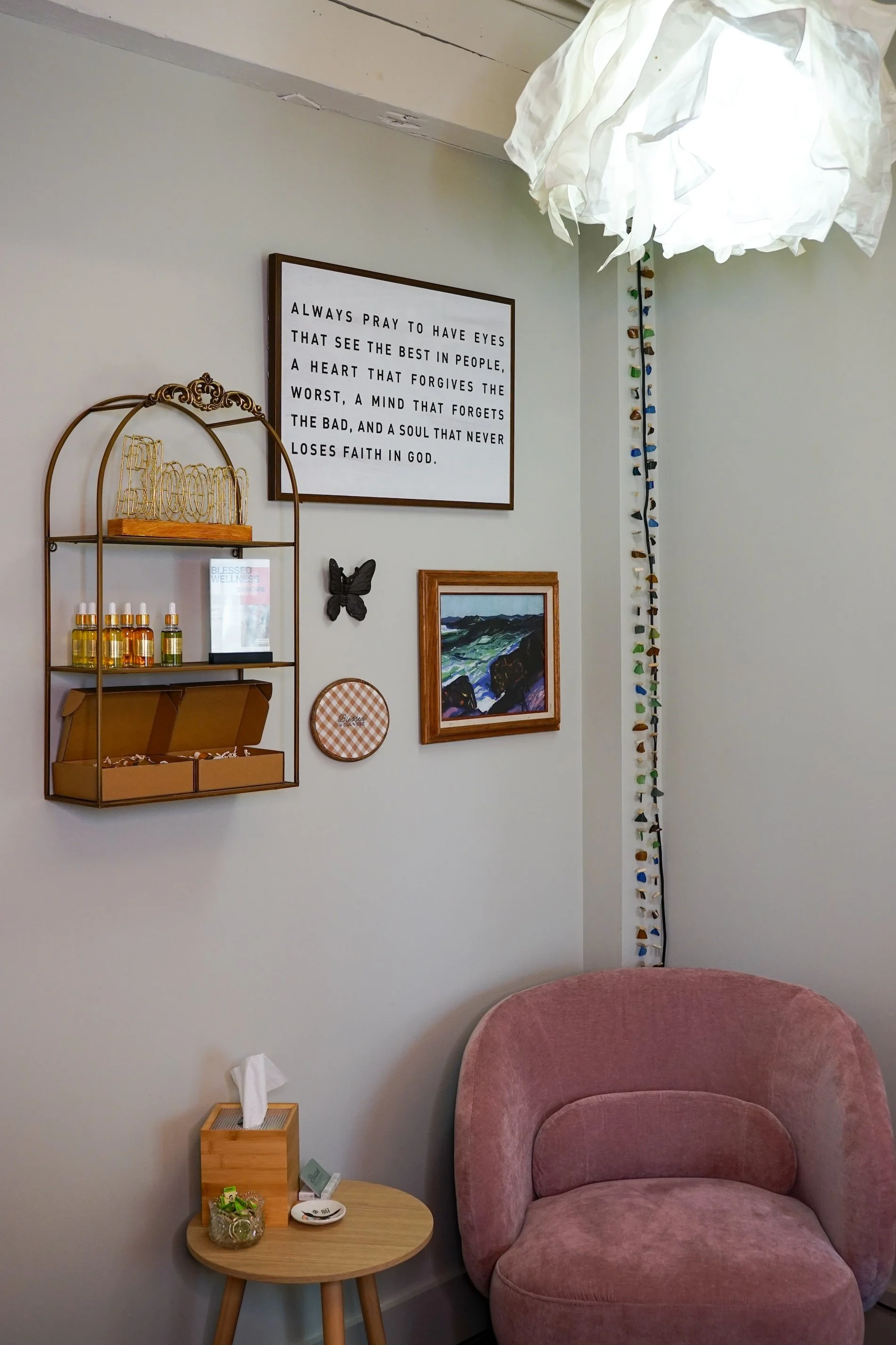 A cozy corner with a pink upholstered armchair, a small circular wooden side table with a tissue box, some small items, and a jar of candies. The wall behind has a framed quote about prayer and faith, a decorative butterfly, and a piece of artwork. There is a hanging light fixture with a crumpled white paper lampshade, and a vertical strip of multicolored ceramic tiles.