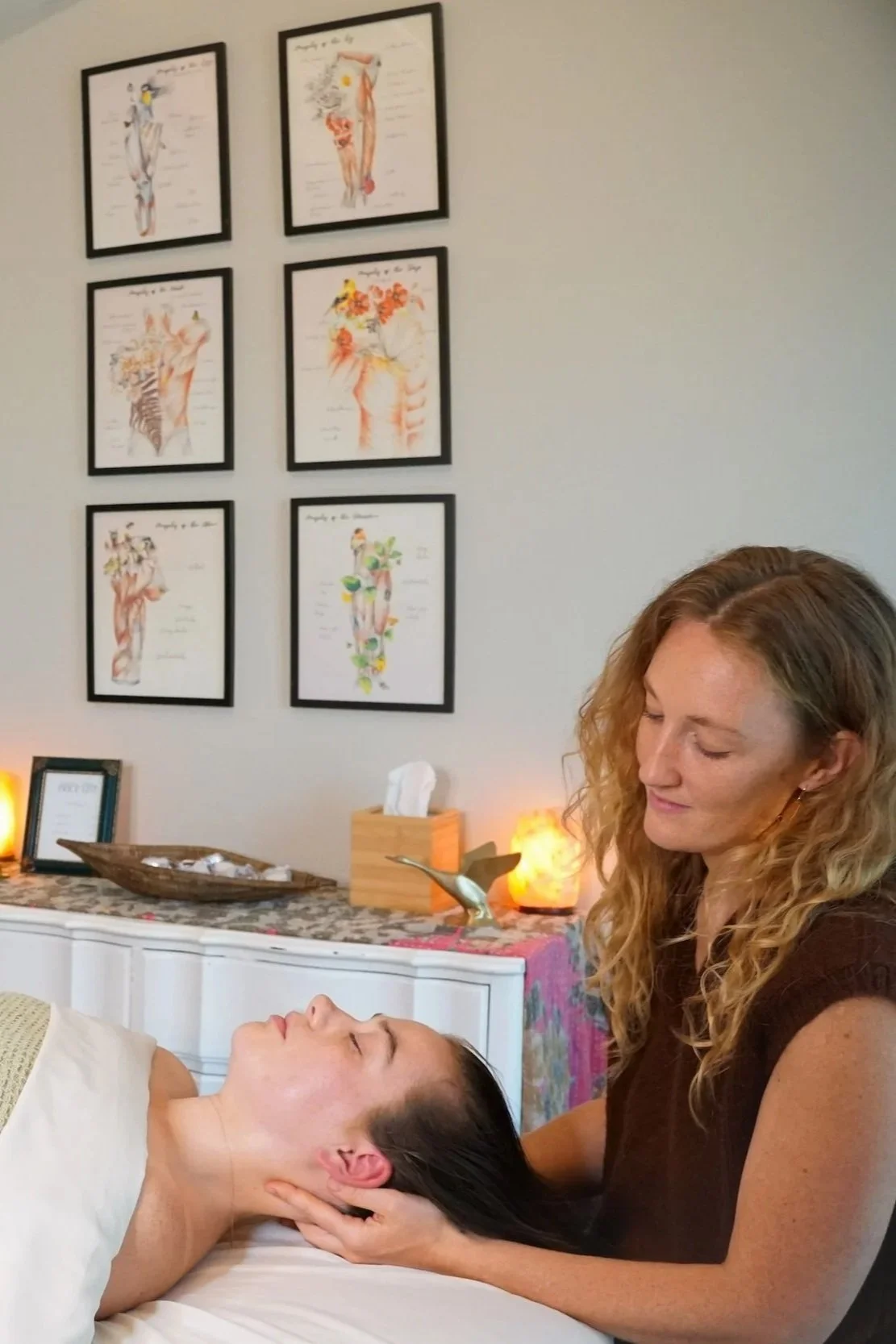 A woman is receiving a massage in a wellness or spa setting, with a massage therapist gently holding her neck. The background features framed anatomical art on the wall and a decorated table with candles, a tissue box, and other spa items.