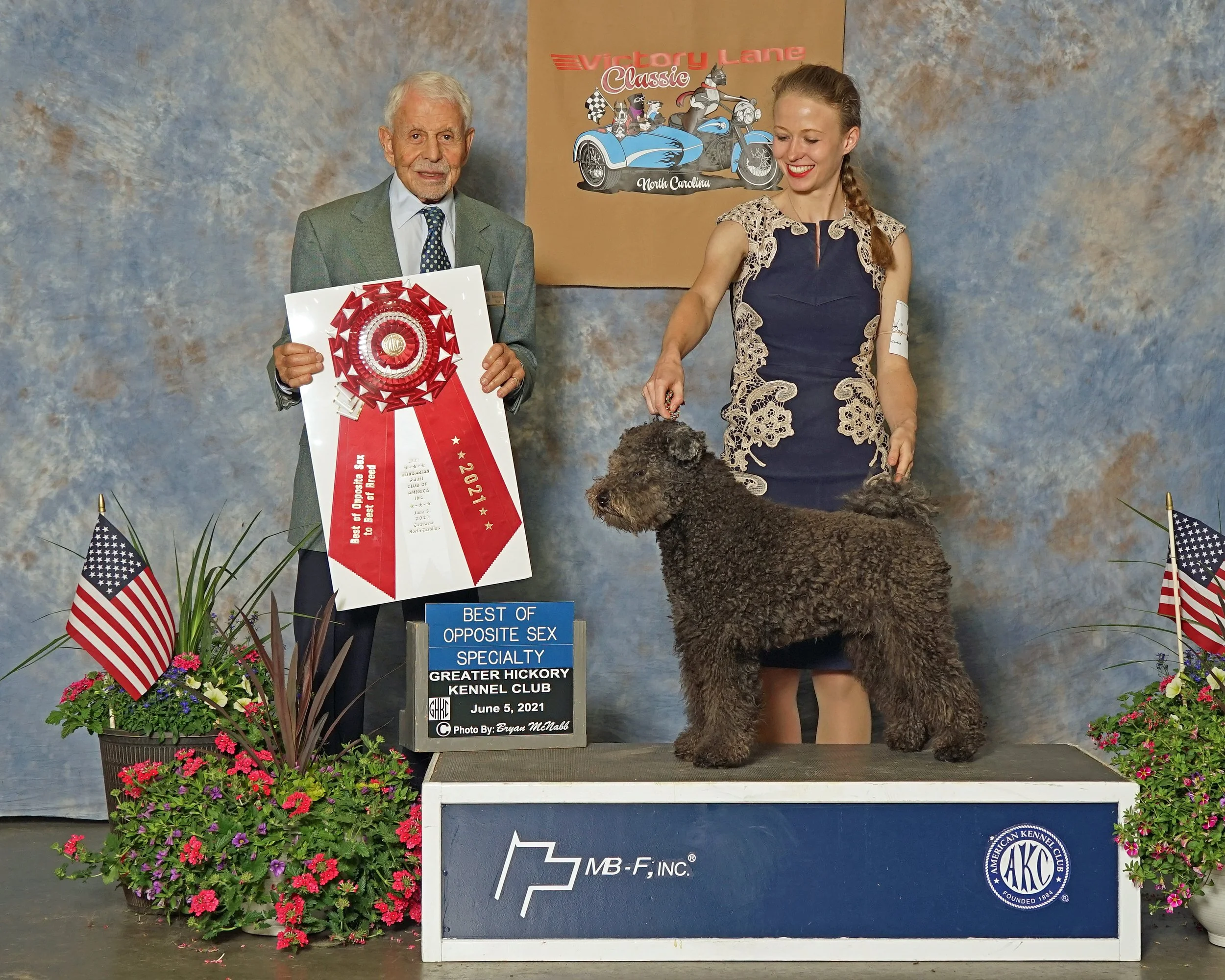 A man and a woman stand behind a grey Hungarian Pumi show dog on a platform. The man holds a large red an white ribbon that says Best of Opposite Sex Specialty. 