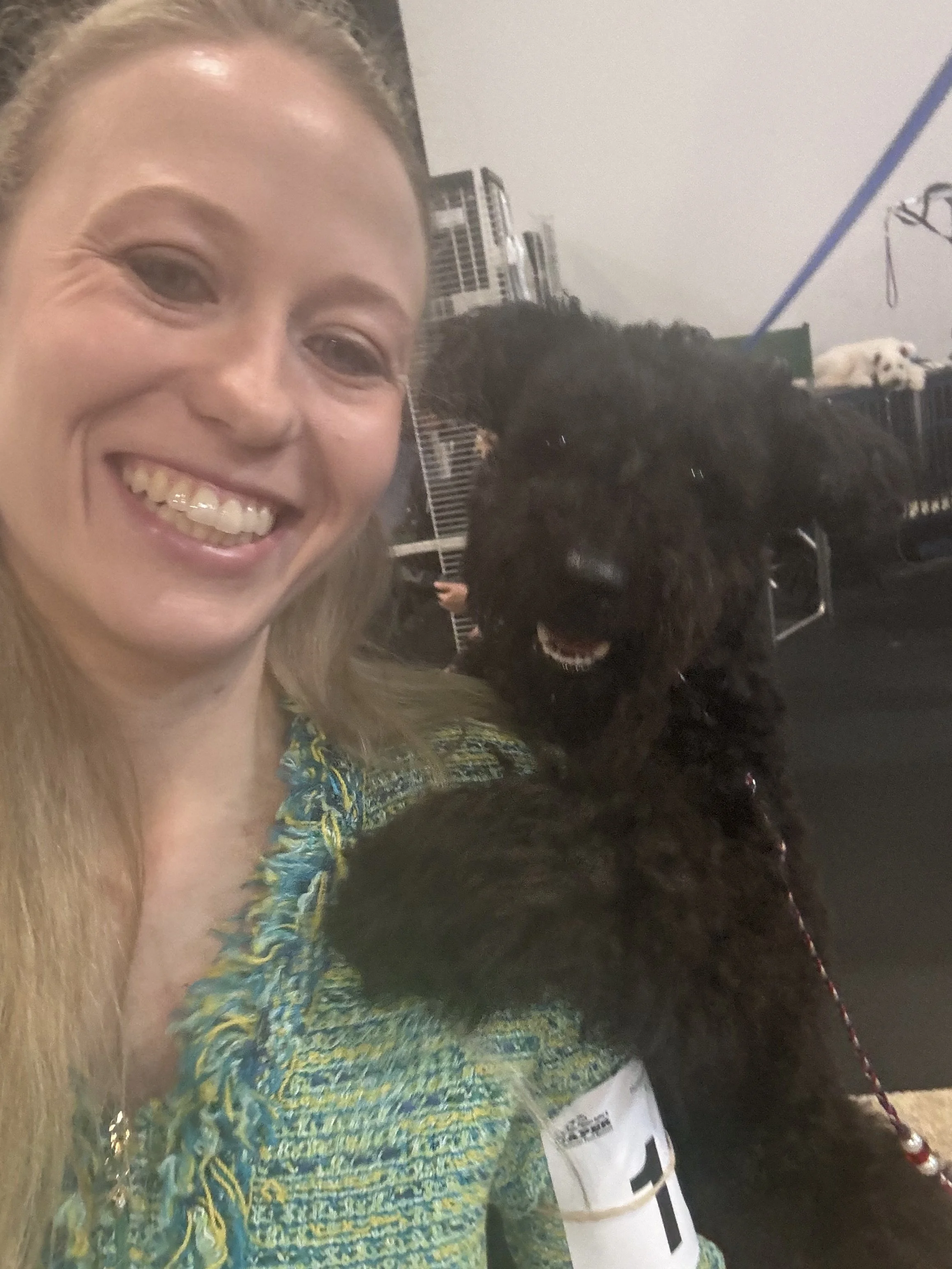 A woman with long blonde hair smiling for a selfie, holding a small black Hungarian Pumi dog with curly fur and a leash, in an indoor setting with crates.