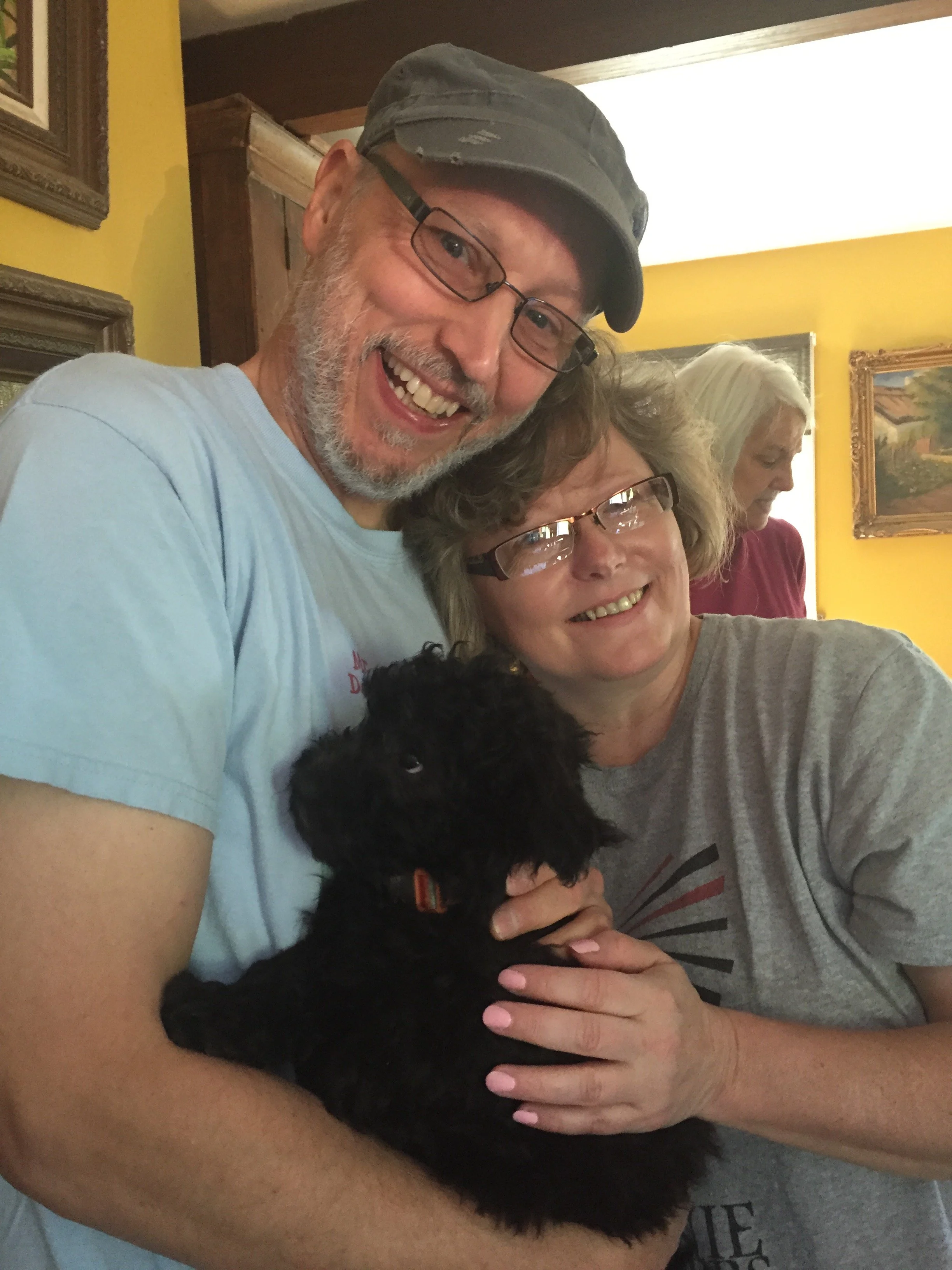 A man and a woman smiling as they hold a small black Pumi puppy named Frida together in a room with yellow walls and framed pictures.