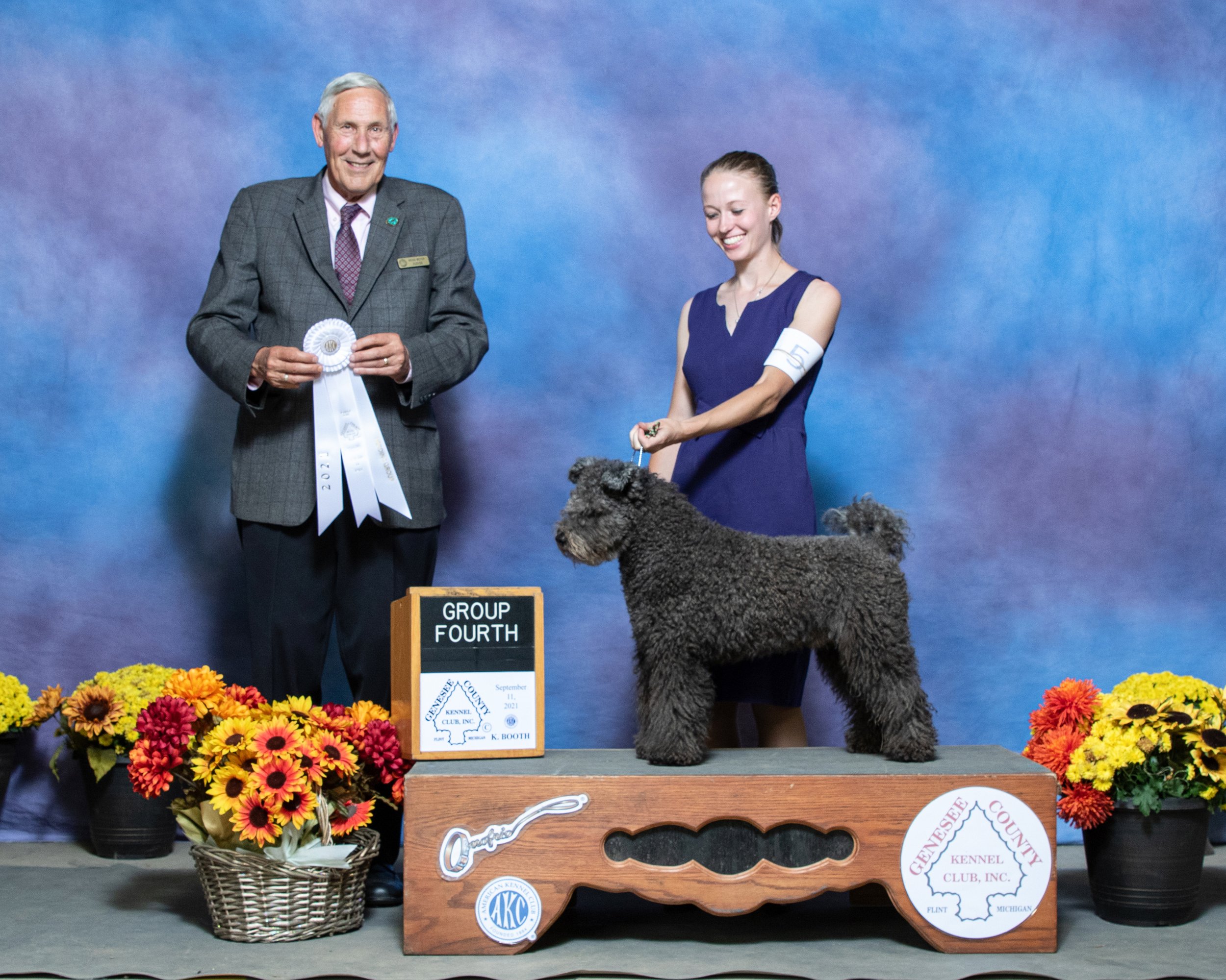 A man in a suit holding a ribbon and a woman in a purple dress with a number on her arm standing on a platform with a black Pumi at a dog show. There are colorful flowers at the front and a sign with the text 'Group Fourth' and other details.