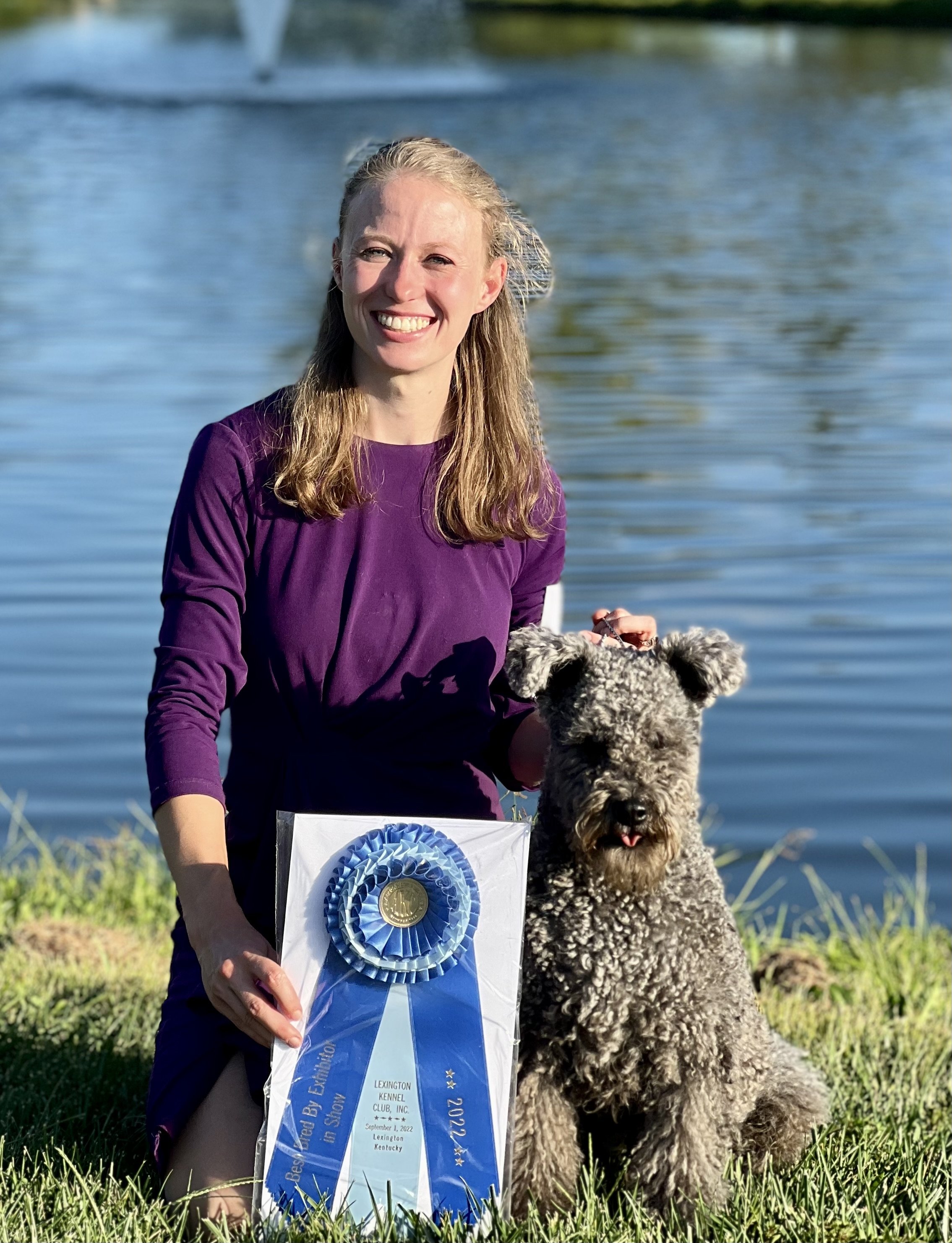 A woman in a purple dress holding a blue ribbon, standing beside a curly-haired Hungarian pumi dog by a lake in the background.