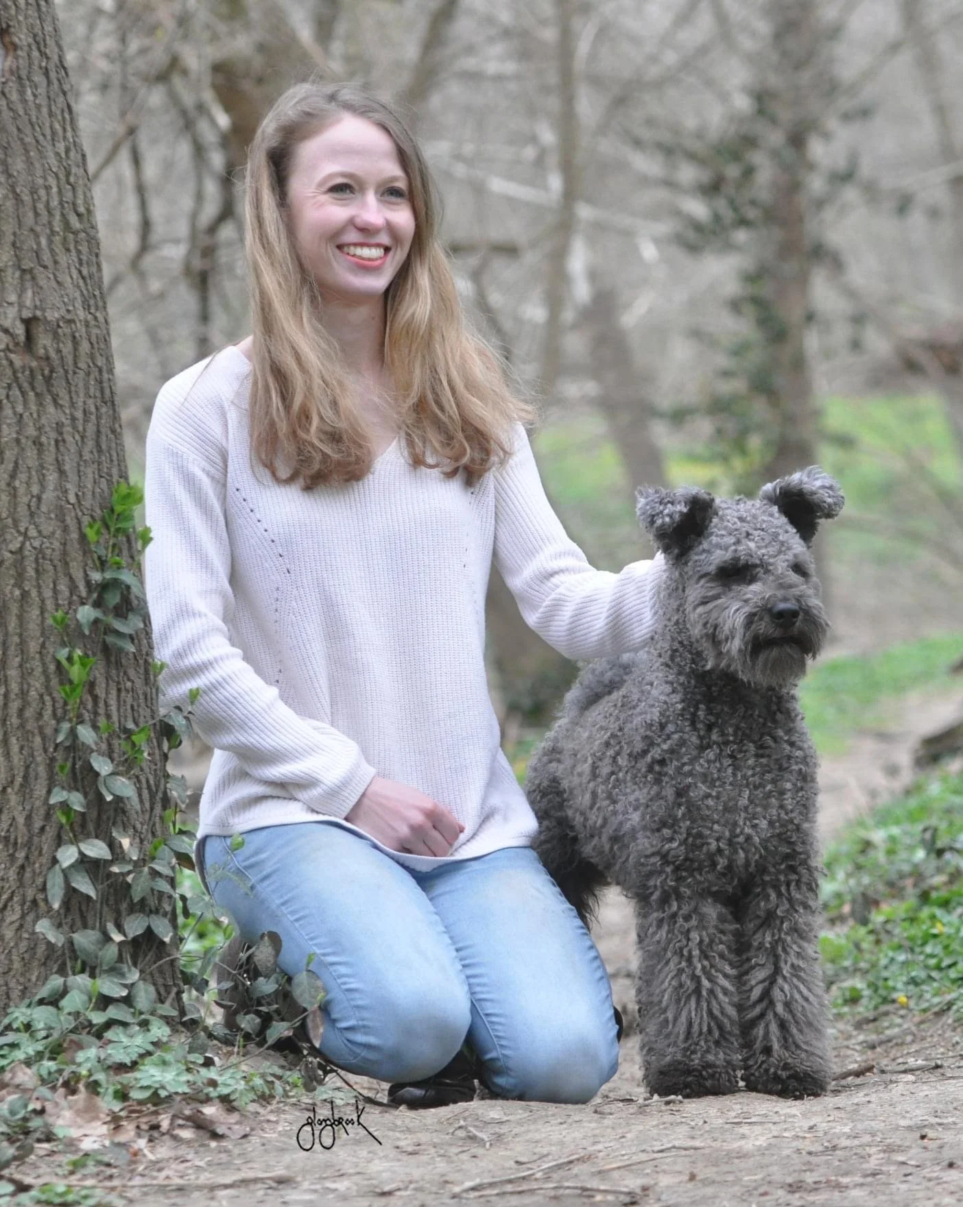 A woman with long blonde hair and a white sweater is kneeling beside her curly-haired gray hungarian Pumi dog outdoors in a wooded area. The woman is smiling and resting her hand on the dog's head, and the dog is sitting on the ground facing forward.