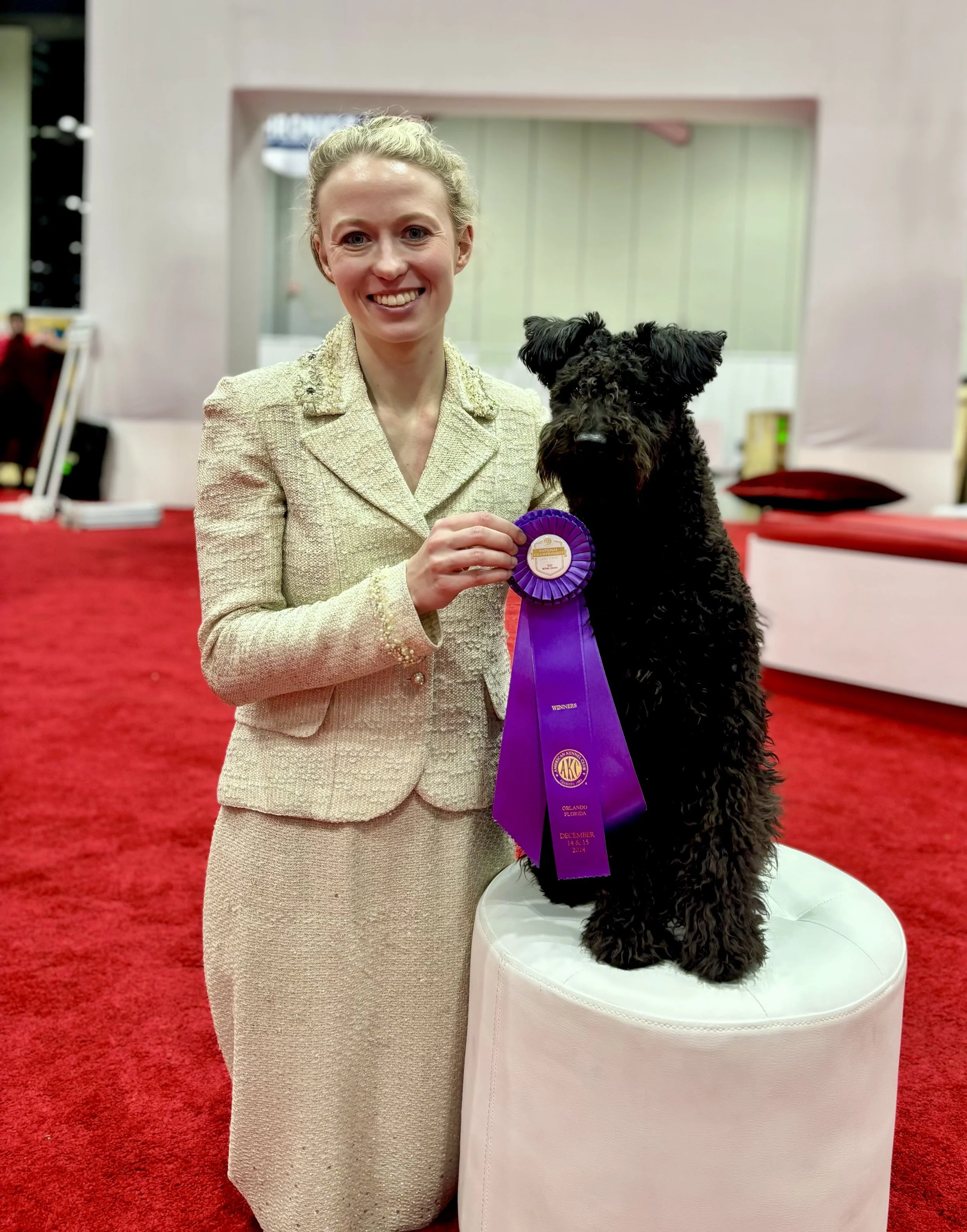 A woman with blonde hair, wearing a beige suit, holding a purple ribbon rosette, standing next to a black Hungarian pumi on a white pedestal, in a room with red carpet and a pink structure in the background.