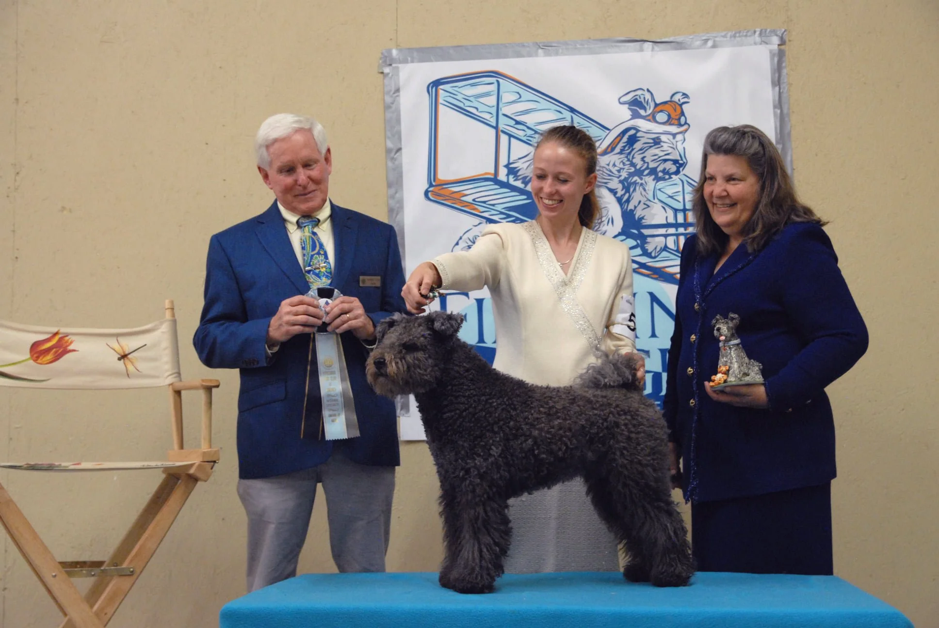 A Hungarian Pumi show dog on a platform, with three people looking down and smiling. The man holds a ribbon. 