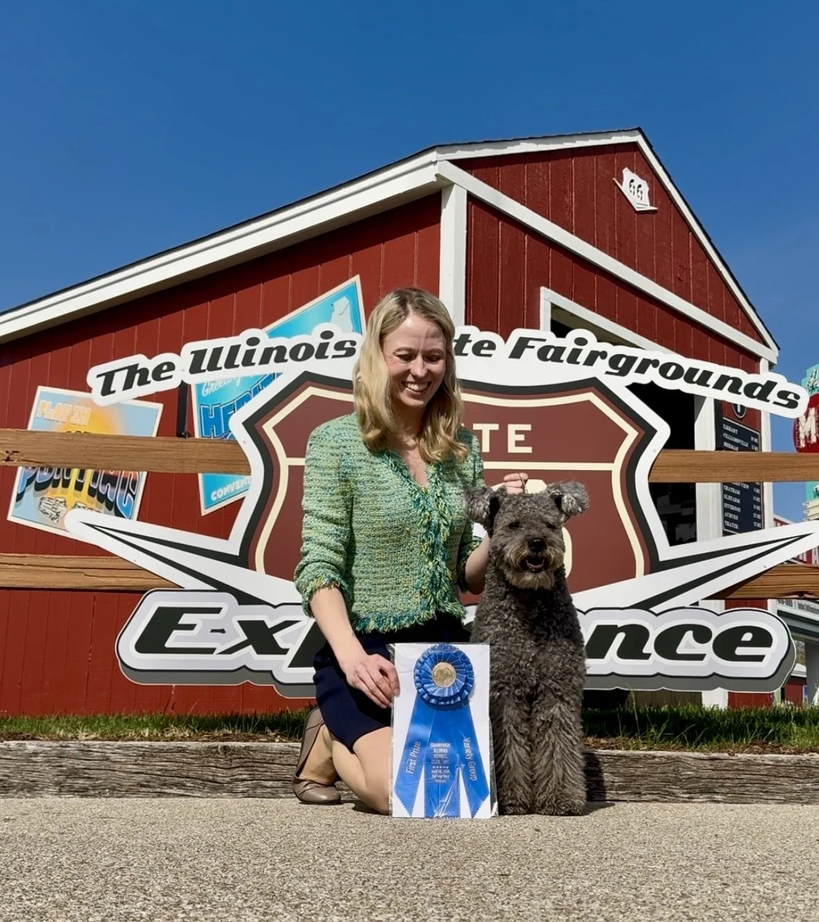 A woman kneeling next to a gray Hungarian Pumi dog holding a blue award ribbon in front of a sign for the Illinois State Fairgrounds.