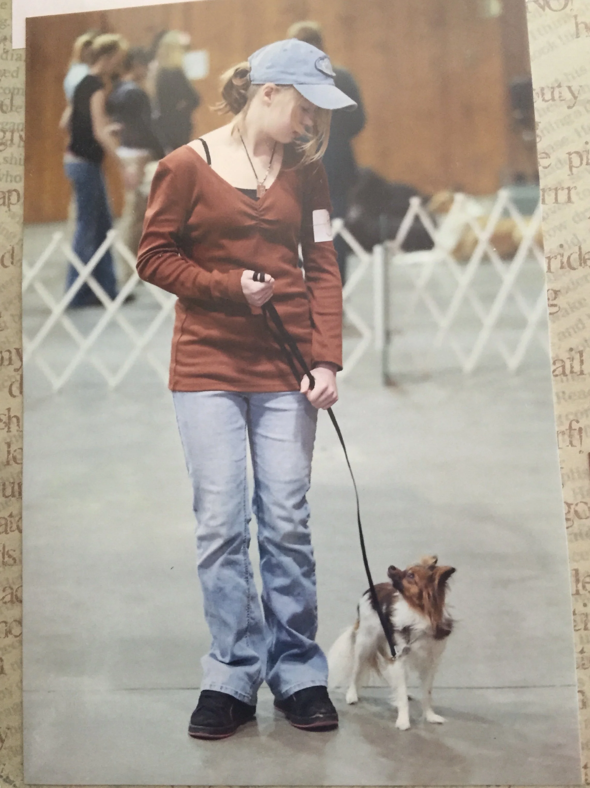 A young woman wearing a baseball cap, red long-sleeve shirt, and light jeans stands holding a leash attached to a small Papillon dog, with others in line in the background.