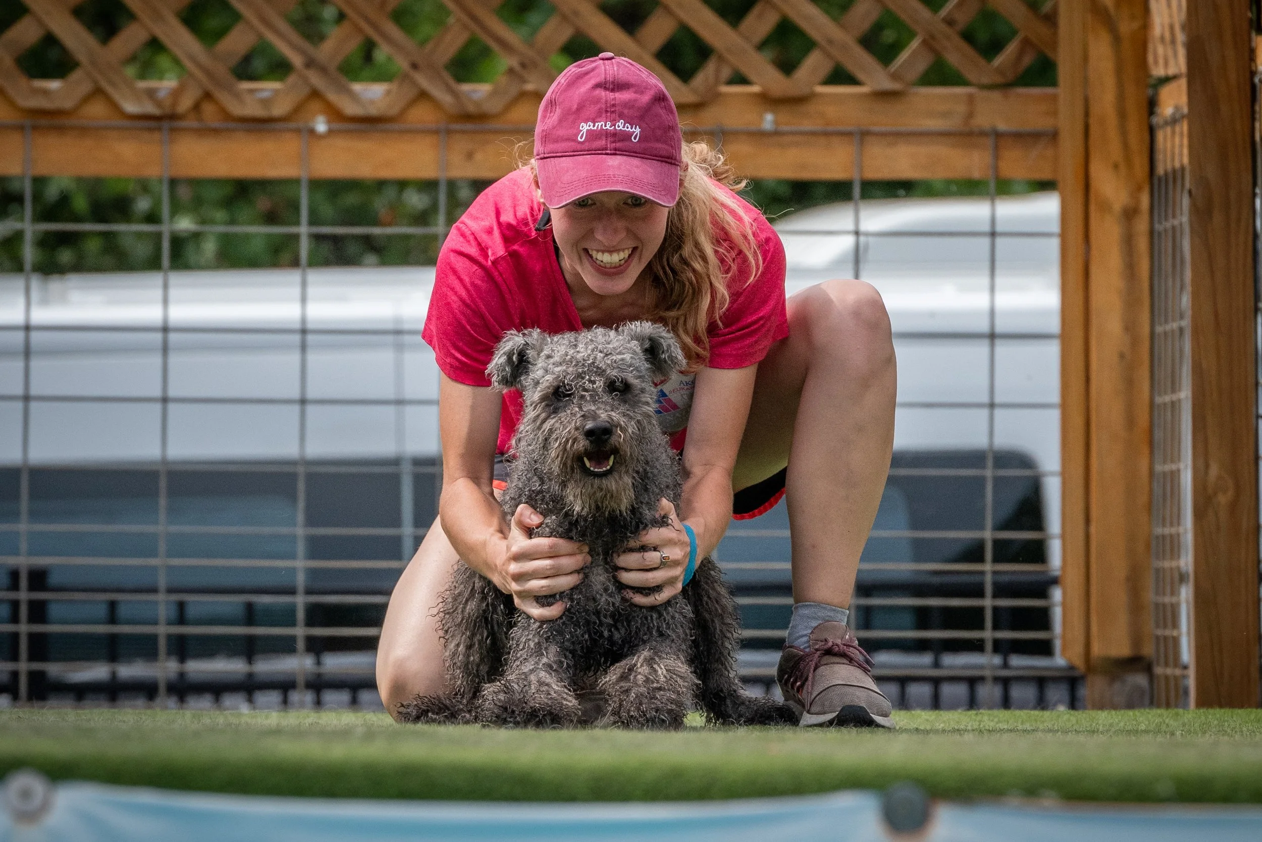 A woman wearing a pink cap and a red shirt is smiling while holding a large curly-haired Hungarian Pumi dog on a grassy field. They are near a wooden and wire fence, with a blurred outdoor background.