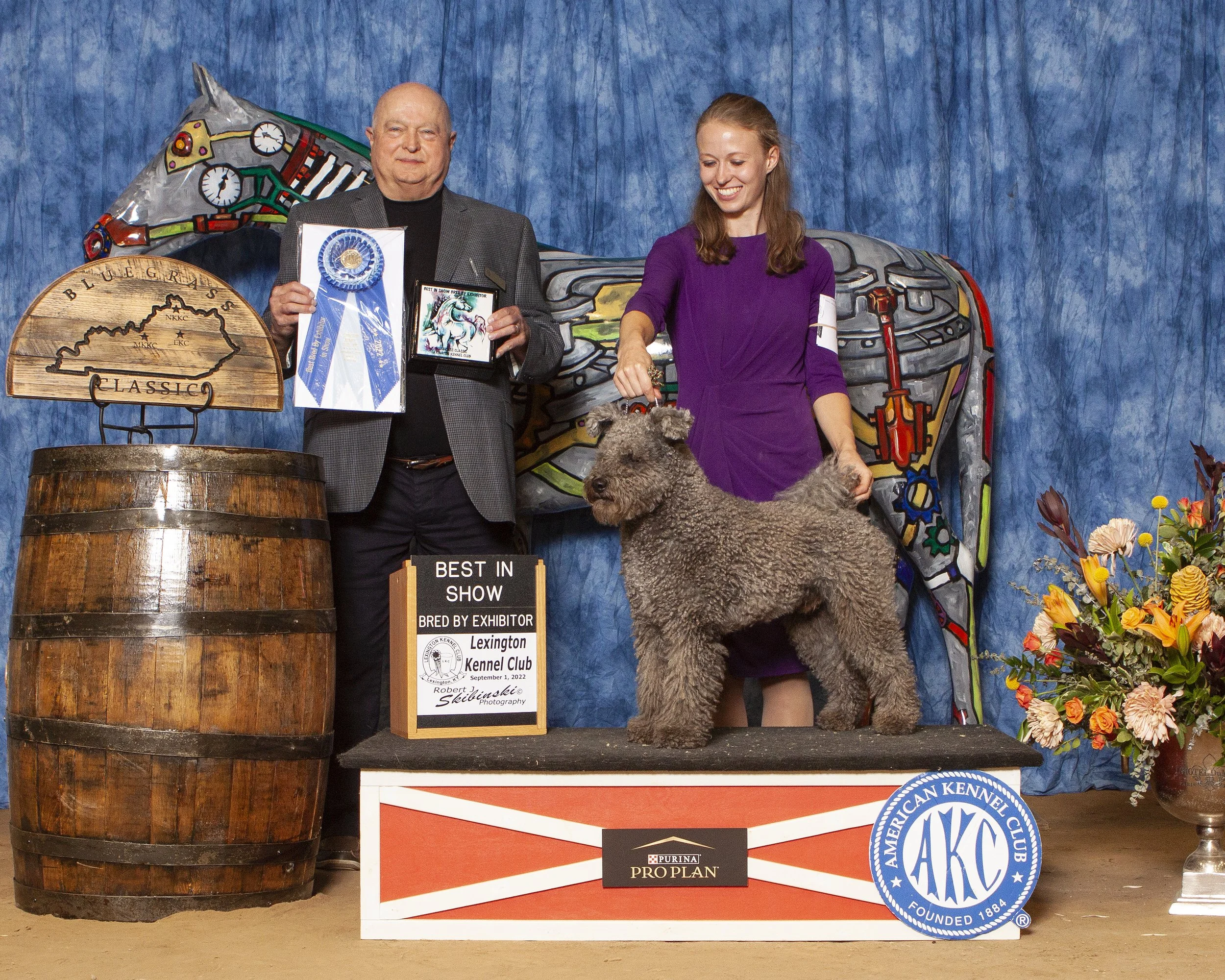 A man and a woman at a dog show, with the man holding a blue ribbon and the woman handling a large, curly-haired Hungarian Pumi dog. There are various awards, signs, and colorful flowers in the background.