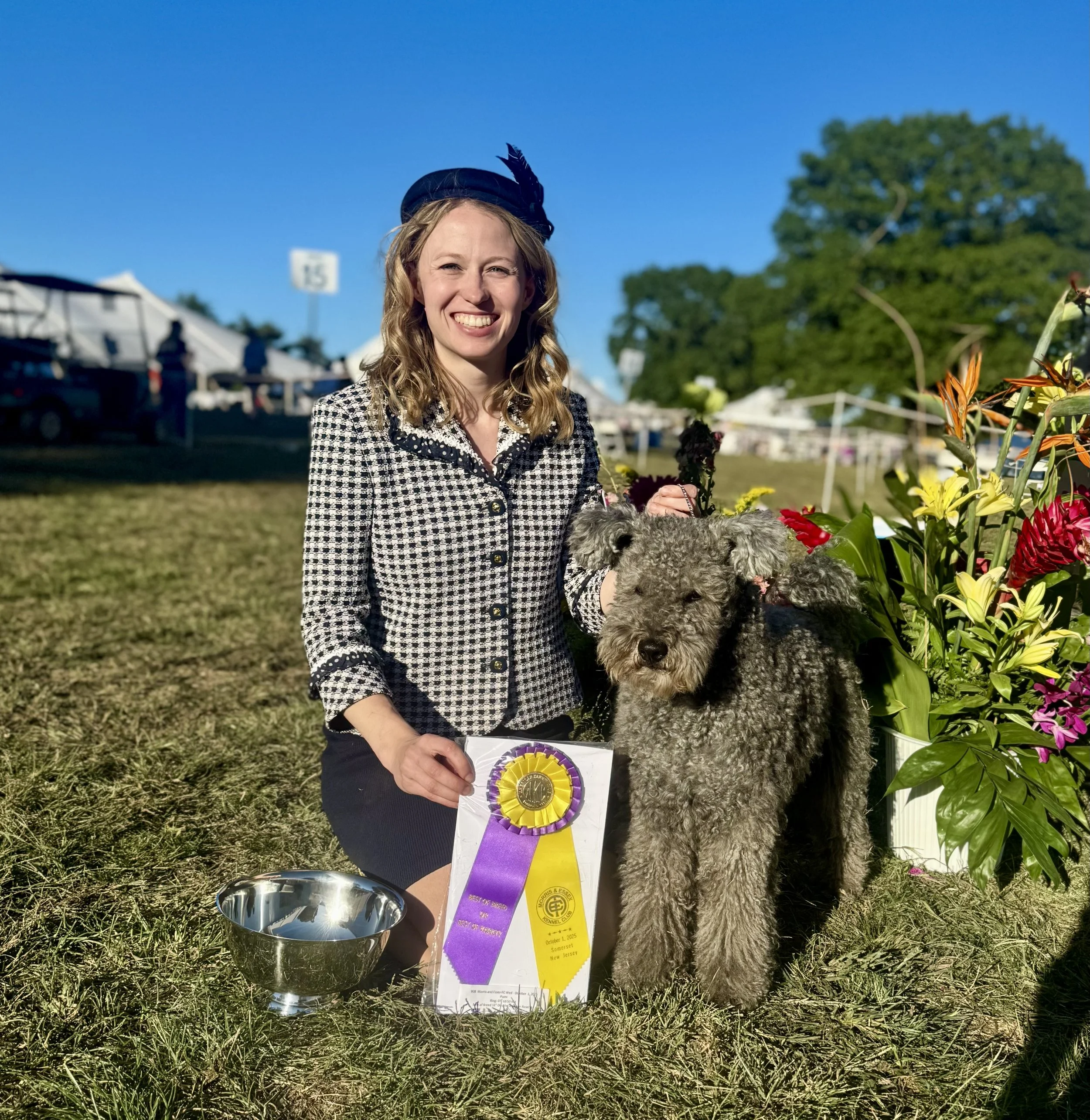 A woman wearing a checkered jacket and a decorative hat is smiling at an outdoor event, holding a silver trophy and a purple and yellow ribbon. She is next to a grey Hungarian Pumi dog. 