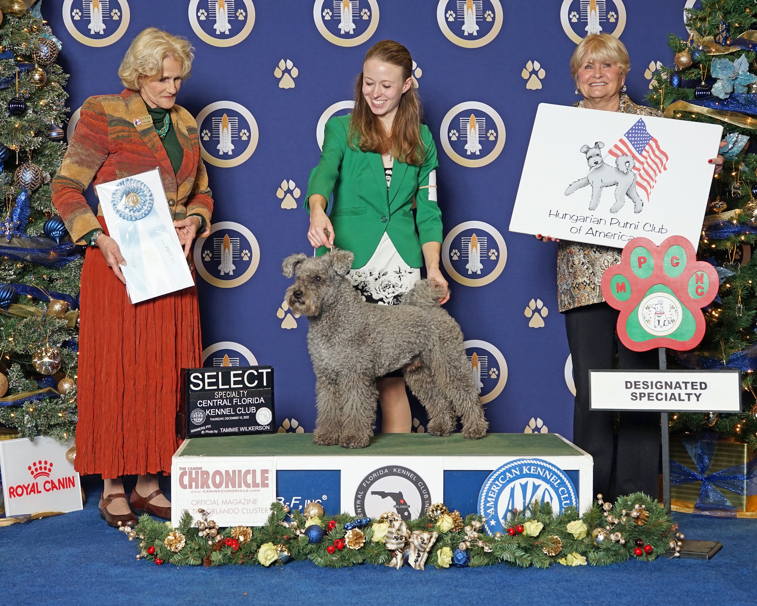 Three women and a dog standing on a platform at a dog show, with Christmas trees decorated with blue and gold ornaments on either side and a blue backdrop featuring logos of a canine organization. The woman in the center is holding a dog, while the w