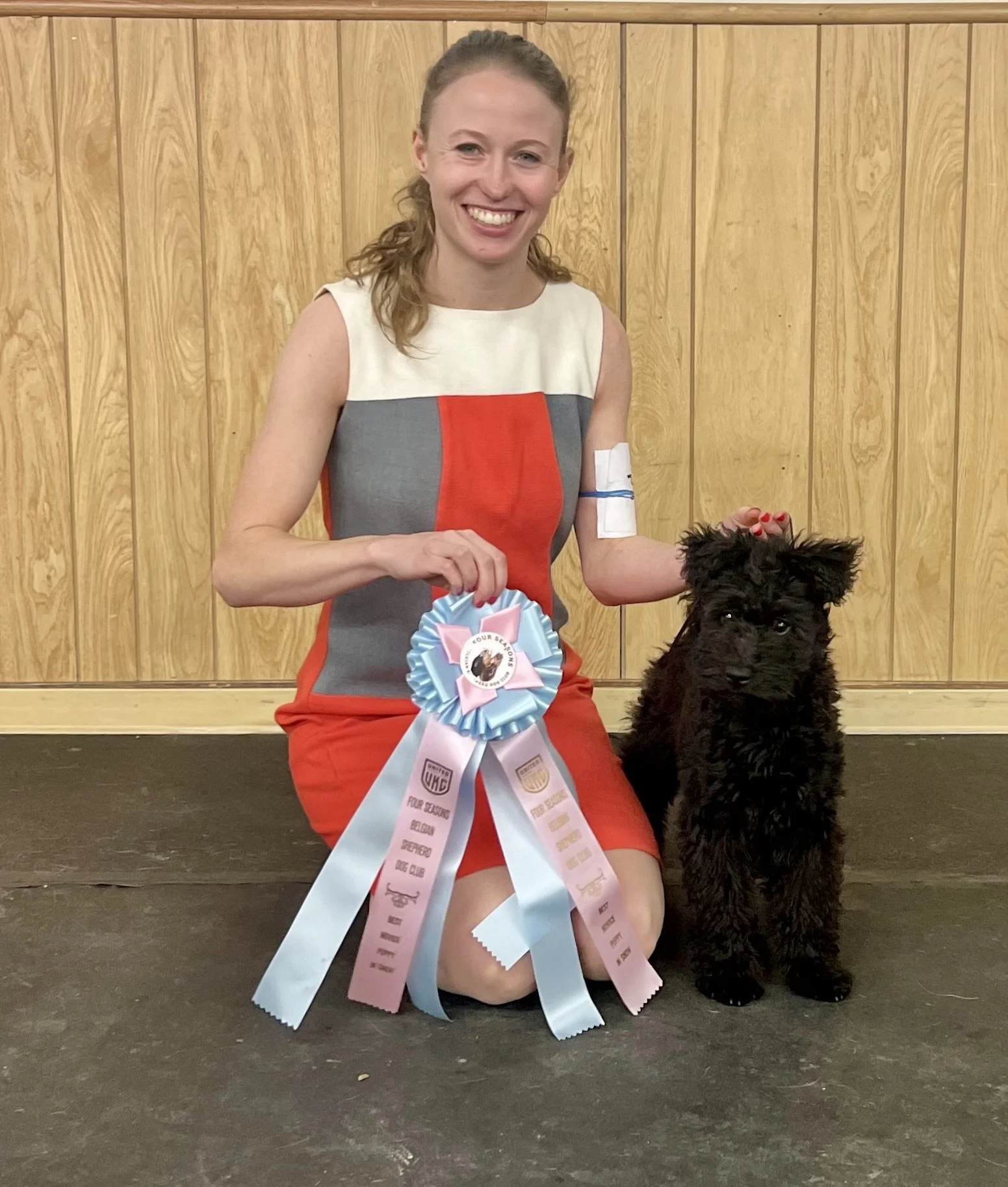 A young woman with red hair tied back, wearing a sleeveless dress with color blocks of white, gray, and red. She is kneeling on one knee, smiling, and holding a large blue and pink ribbon award. Next to her is a black Hungarian Pumi puppy. 