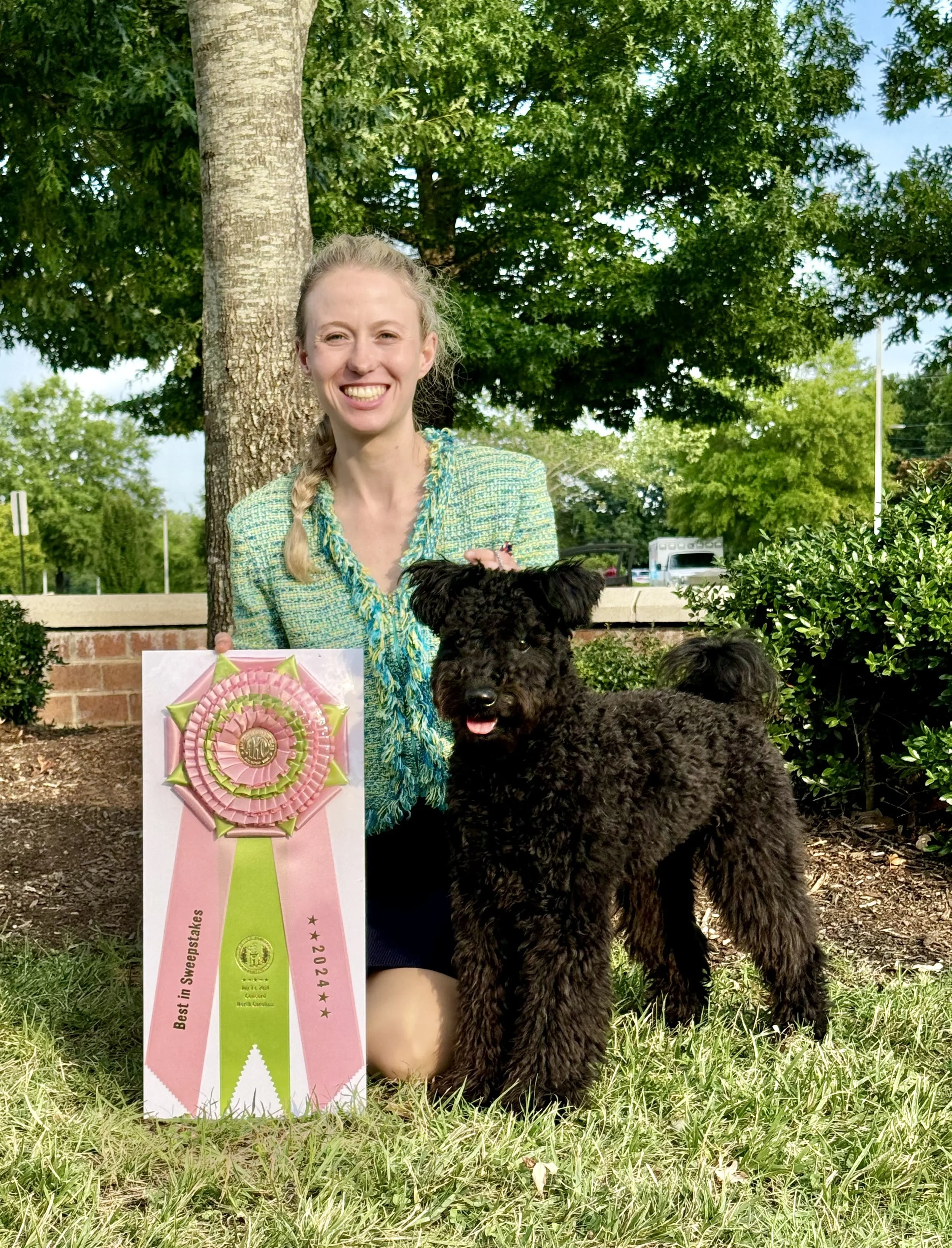 A woman with blonde hair and a braid, smiling, holding a pink award ribbon, with a black Hungarian Pumi dog in front of her, outdoors near a tree and bushes.
