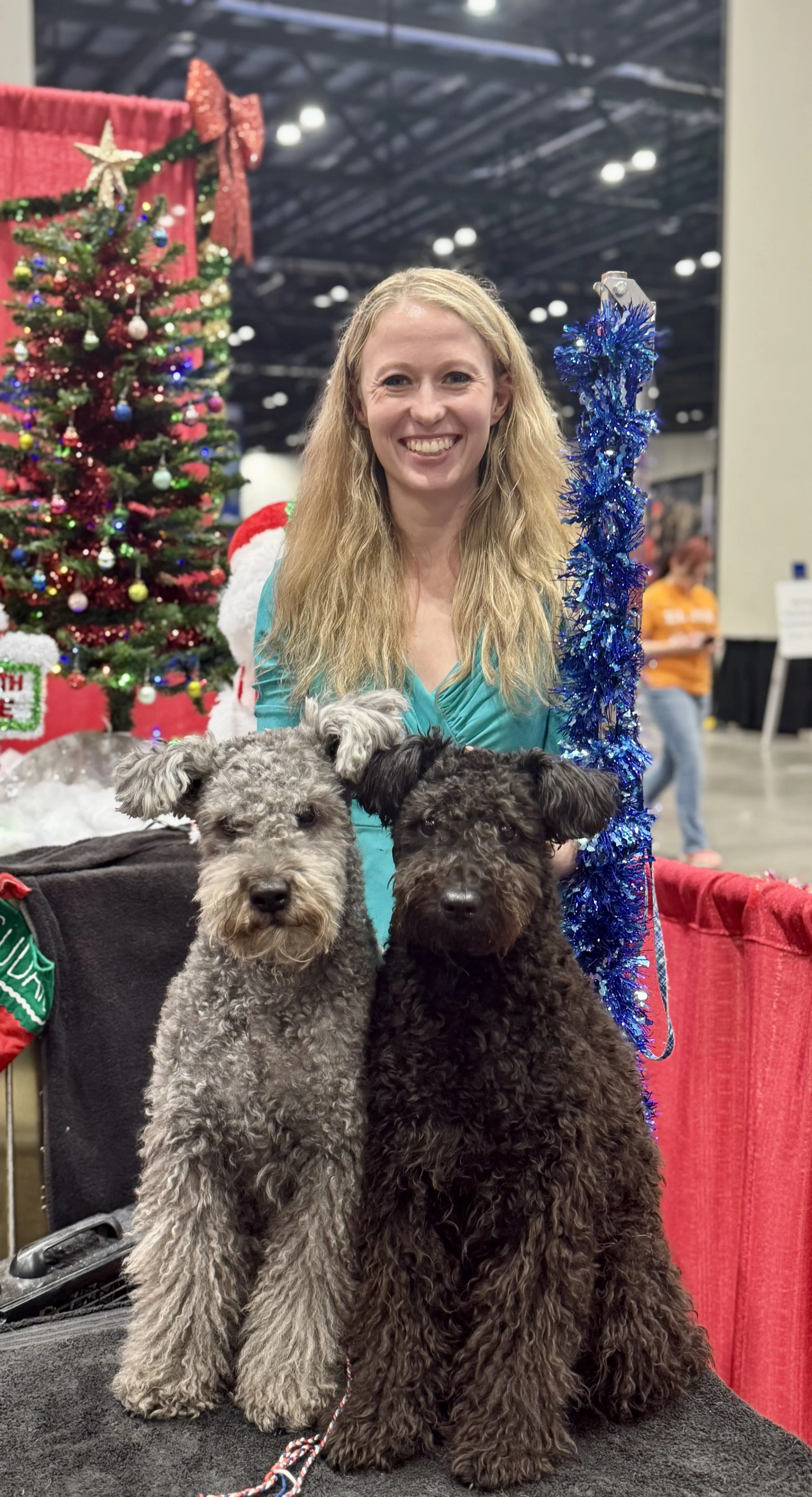 A woman with long blonde hair smiling at the camera, posing with two curly-haired Hungarian Pumi dogs, in front of a decorated Christmas tree at a festive event.