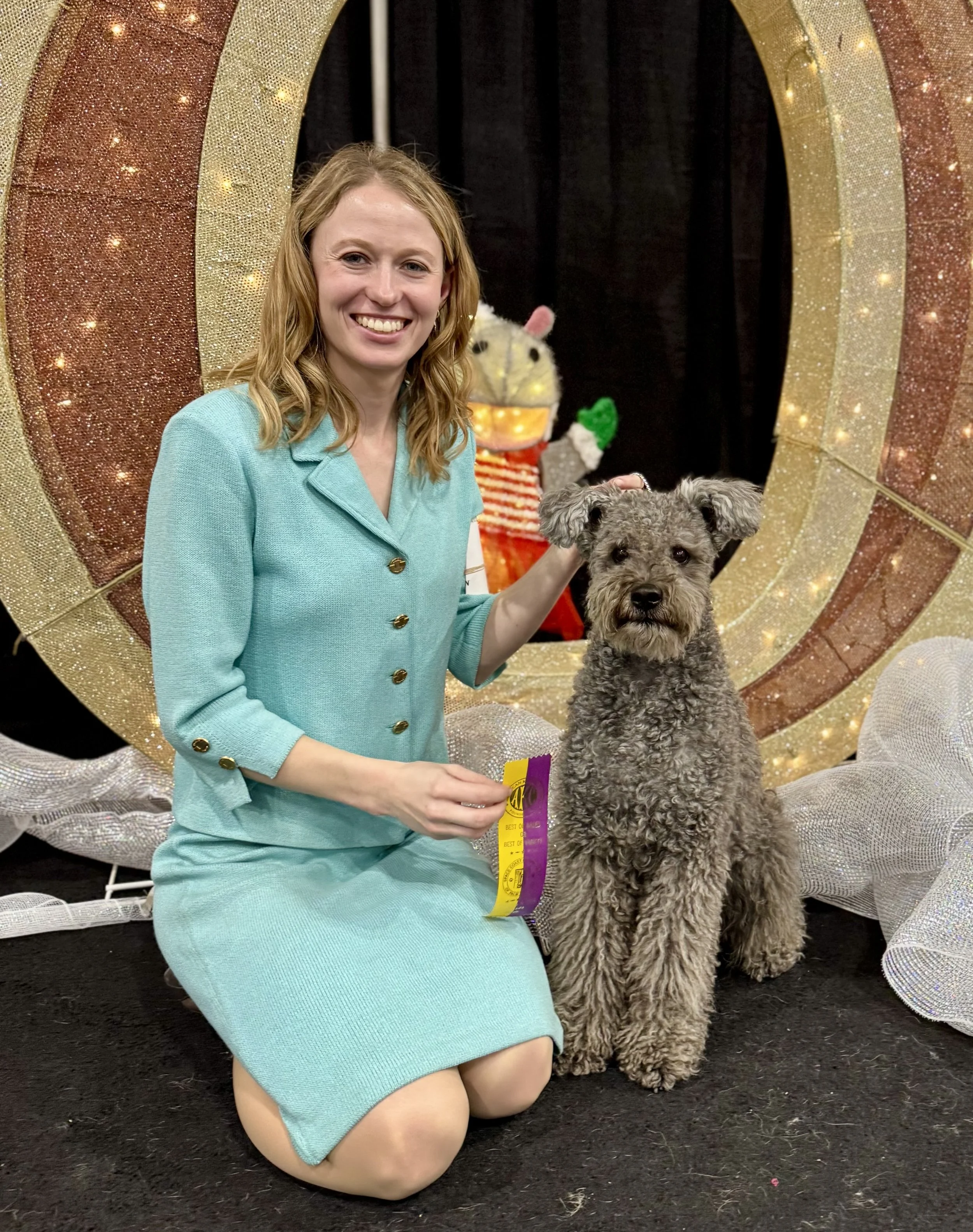 A woman kneeling on a black carpet, smiling, holding a small, curly-haired Hungarian Pumi dog with a yellow and purple ribbon, in front of a sequined gold and bronze circular backdrop, with a plush zebra figure in the background.