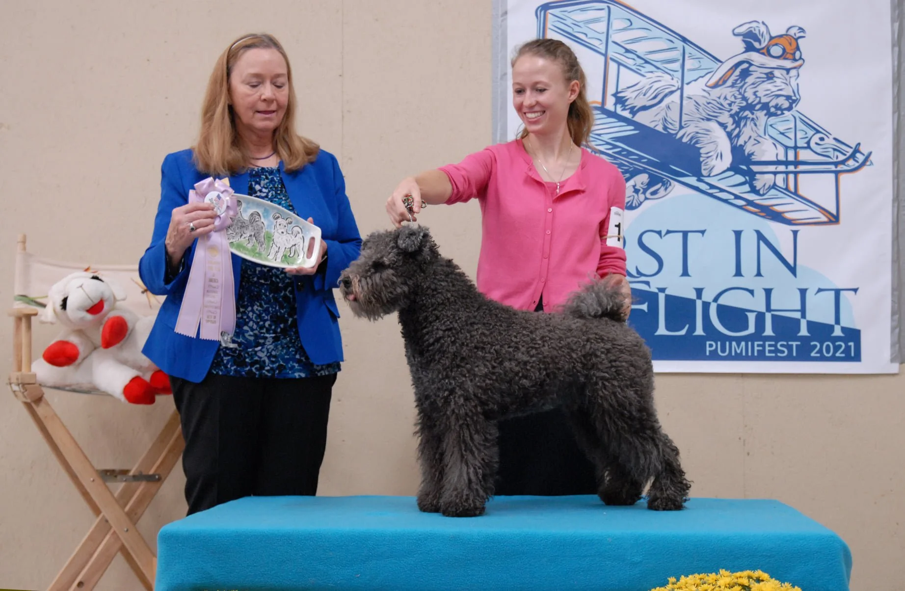 A Hungarian Pumi Dog is on a platform with two women looking down and smiling. One woman holds a ribbon.