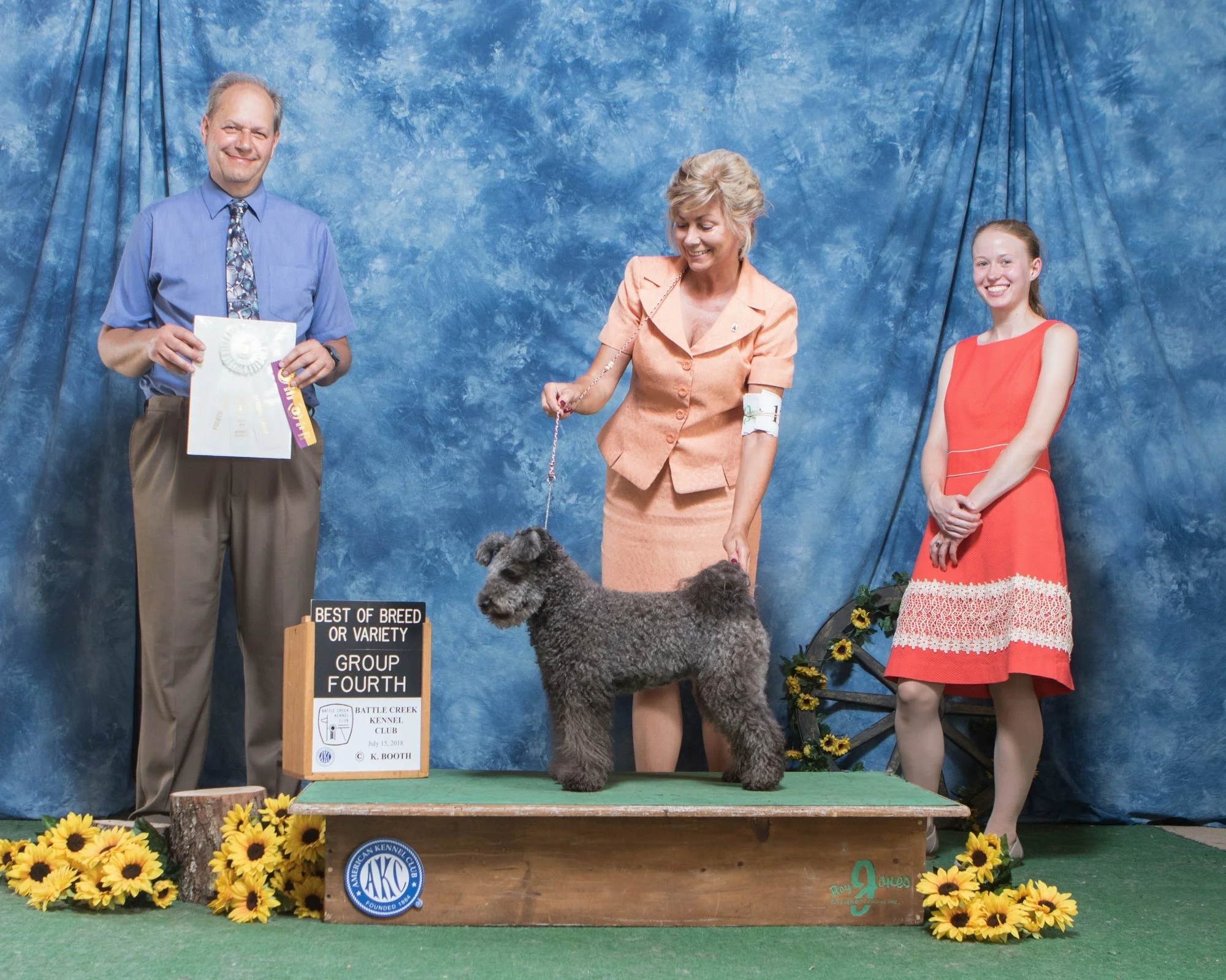 Three people and a dog at a dog show. The man on the left is holding a certificate, the woman in the center is holding a leash attached to a dark gray Hungarian Pumi dog and the woman on the right is smiling. The dog is standing on a green platform.