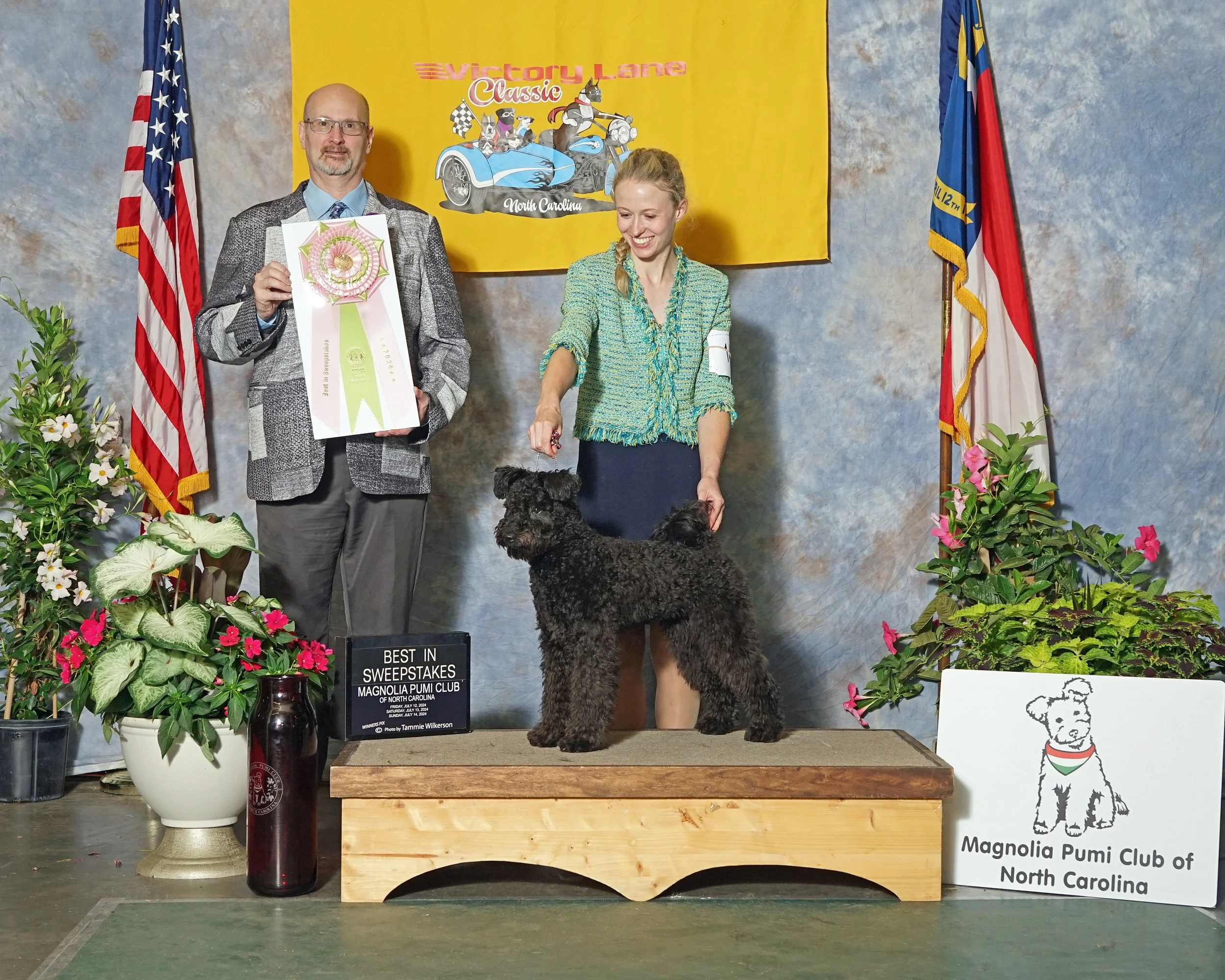 A man, woman, and black Hungarian Pumi dog stand surrounded by flower arrangements and flags. The man holds a ribbon and there is a sign indicating a sweepstakes event. The woman is smiling and looking at the dog, which is standing on the podium.