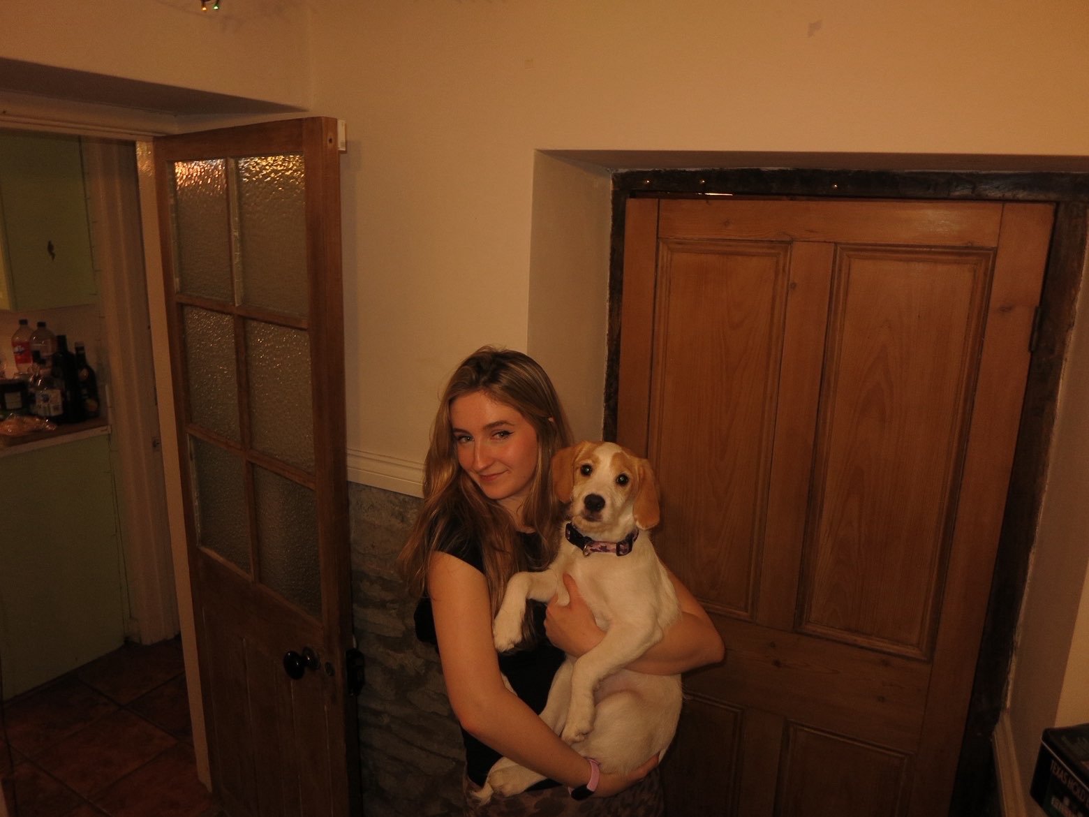 A young woman with long, wavy light brown hair holding a tan and white dog indoors near a wooden door and a half-open glass-paneled door.