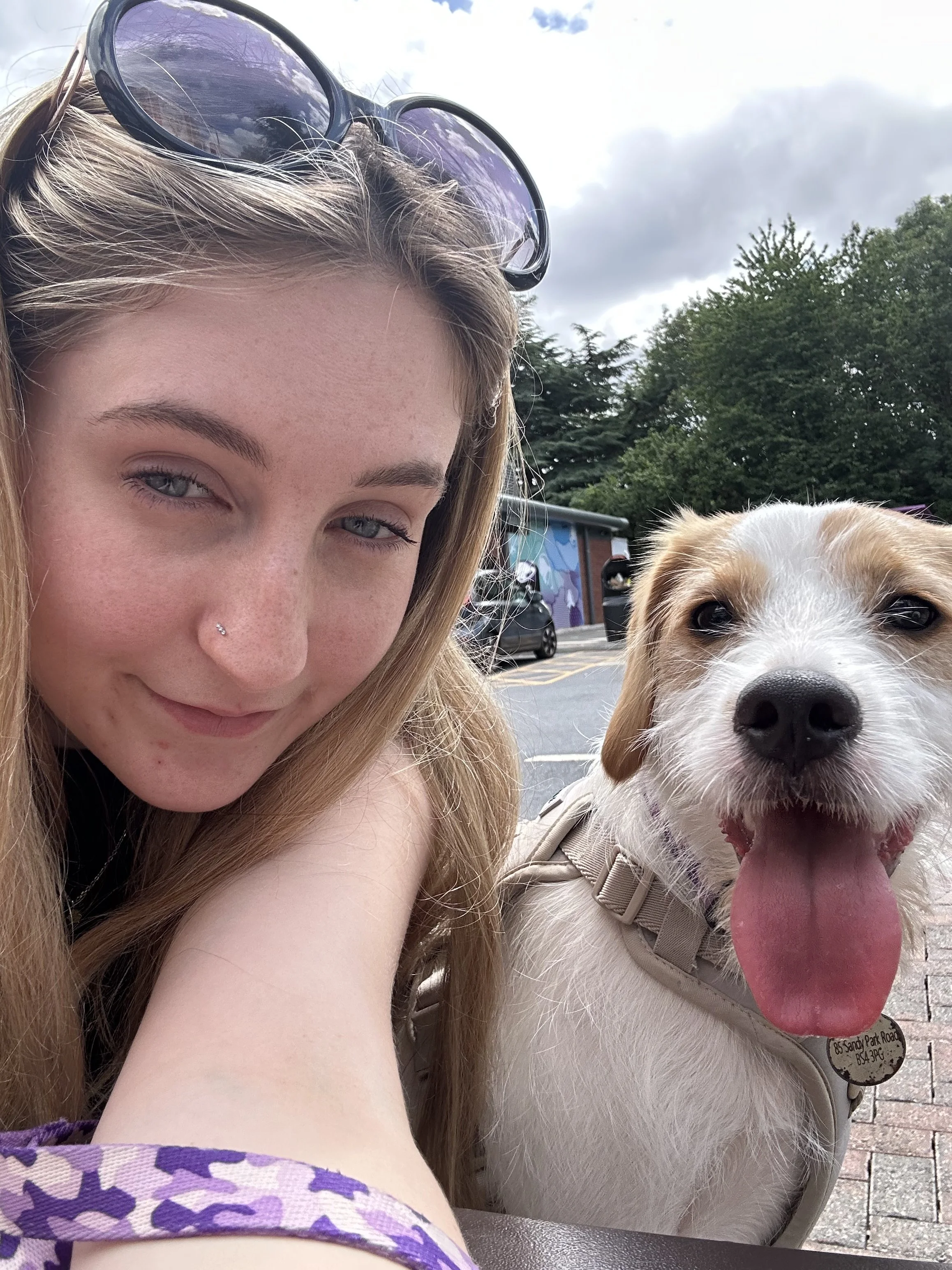 Close-up of a young woman with sunglasses on her head and a nose piercing, smiling next to a happy dog wearing a harness. They are outdoors, with a parking lot and trees in the background.