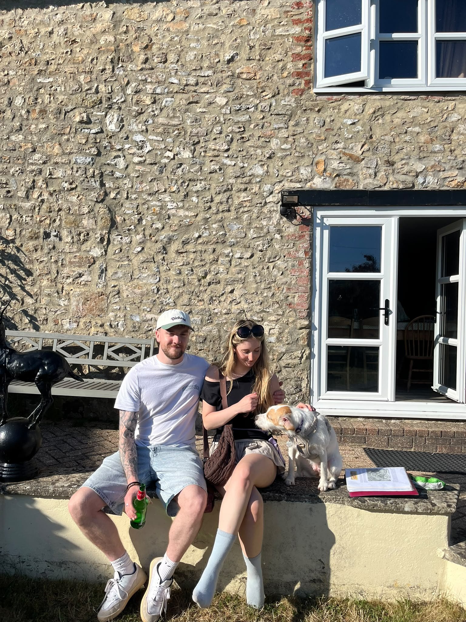 Two young adults, a man and a woman, sitting on a low wall outside a brick house with open windows. The man is holding a green bottle, the woman is petting a white and brown dog, and there are magazines or papers on the ground near them. They appear to be enjoying a sunny day.