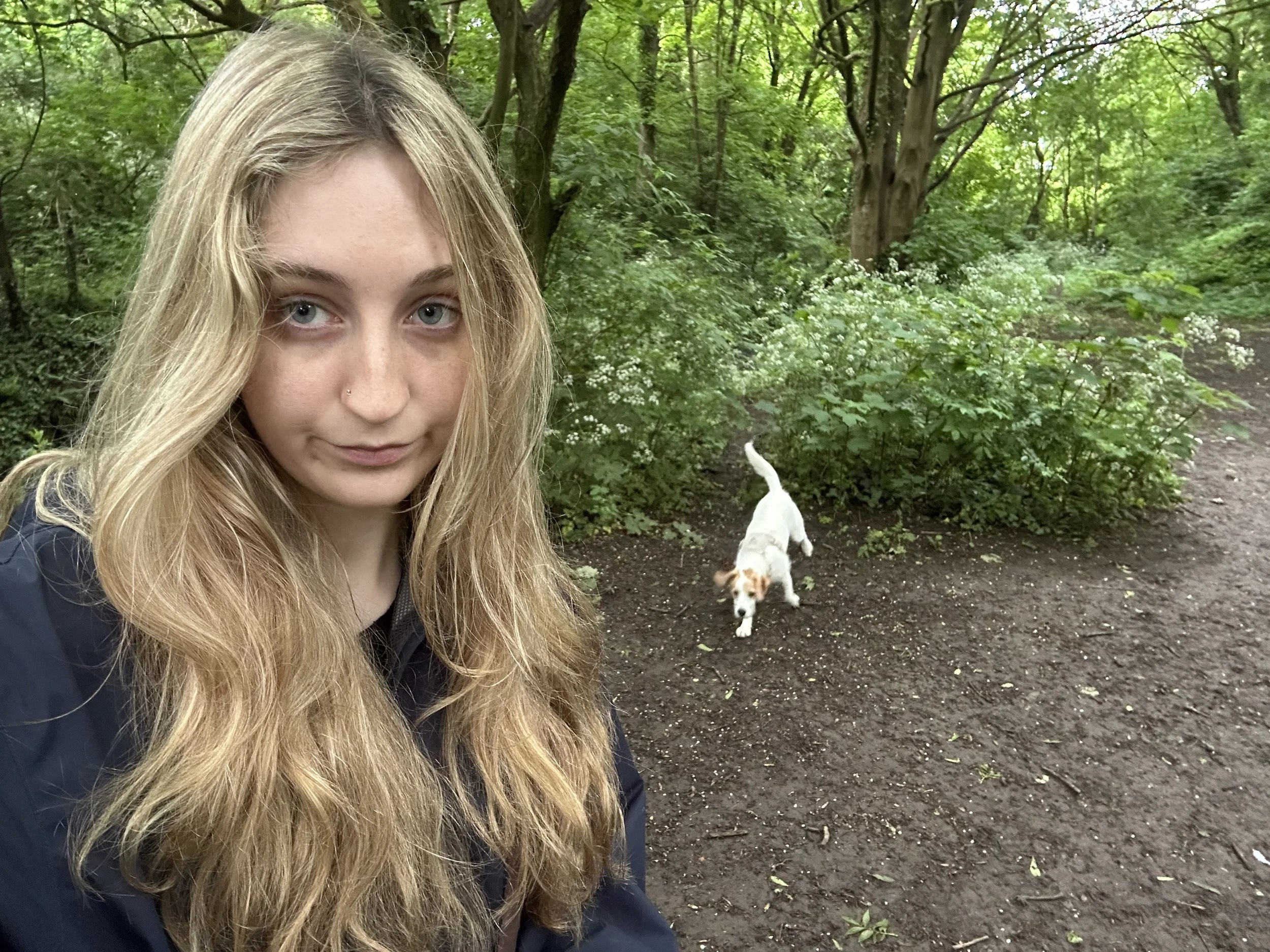 A female dog-trainer with long blonde hair and a small nose piercing taking a selfie in a green wooded area with one dogs, one with a mostly white coat and brown markings, walking on the dirt trail behind her.