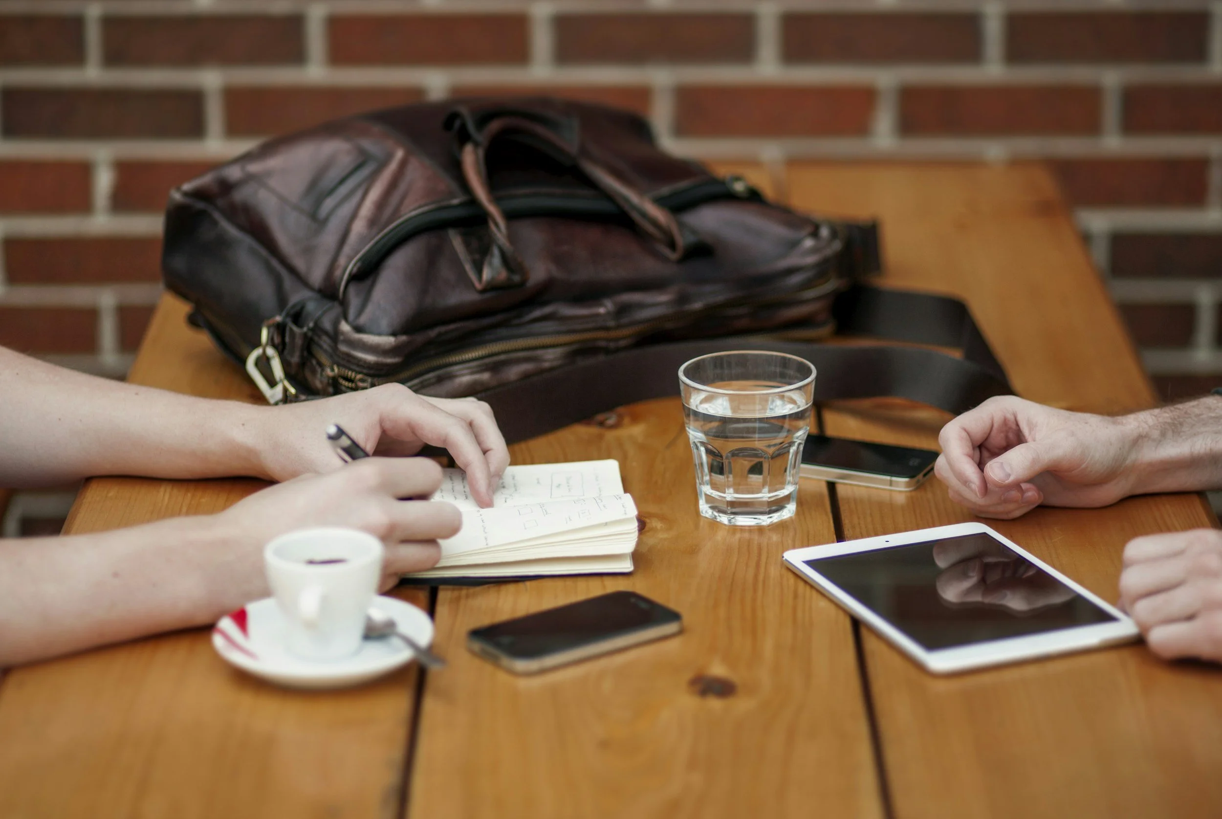 People sitting at a wooden table with notebooks, a glass of water, smartphones, and a coffee cup, against a brick wall.
