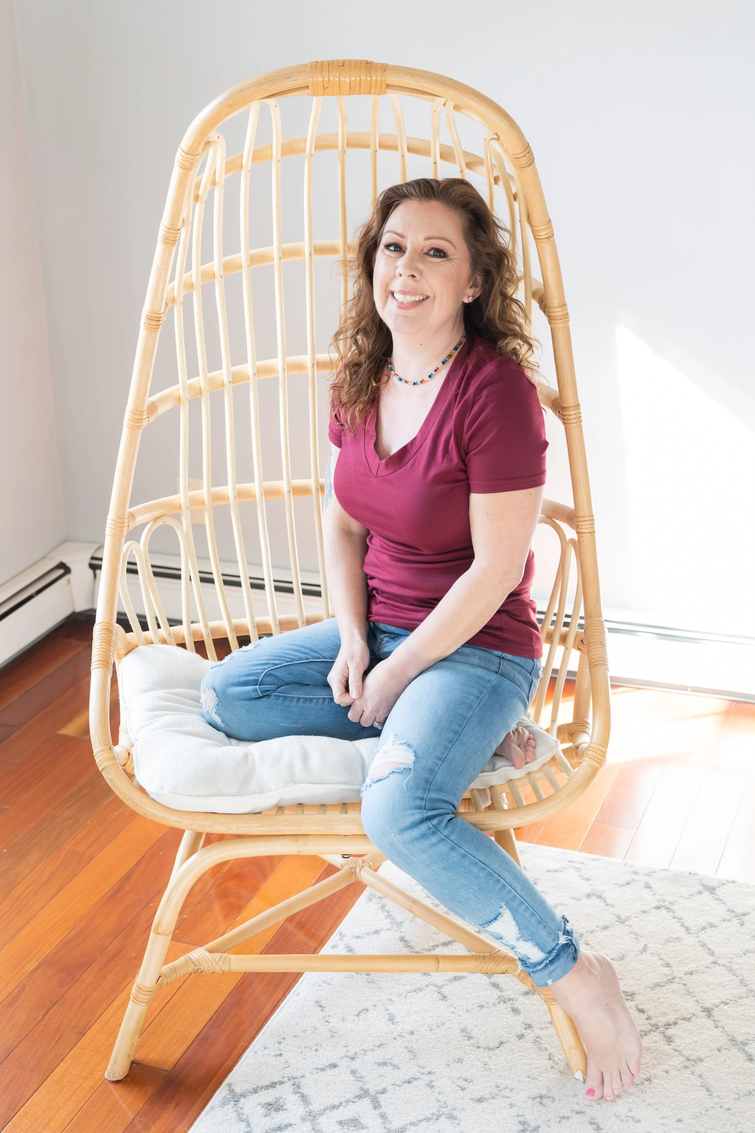 A woman sitting on a round cushioned chair with a high curved back made of rattan, positioned in a living room with hardwood floors and a light-colored rug. She has curly brown hair, is smiling, and is wearing a maroon T-shirt, blue ripped jeans, and a colorful beaded necklace.