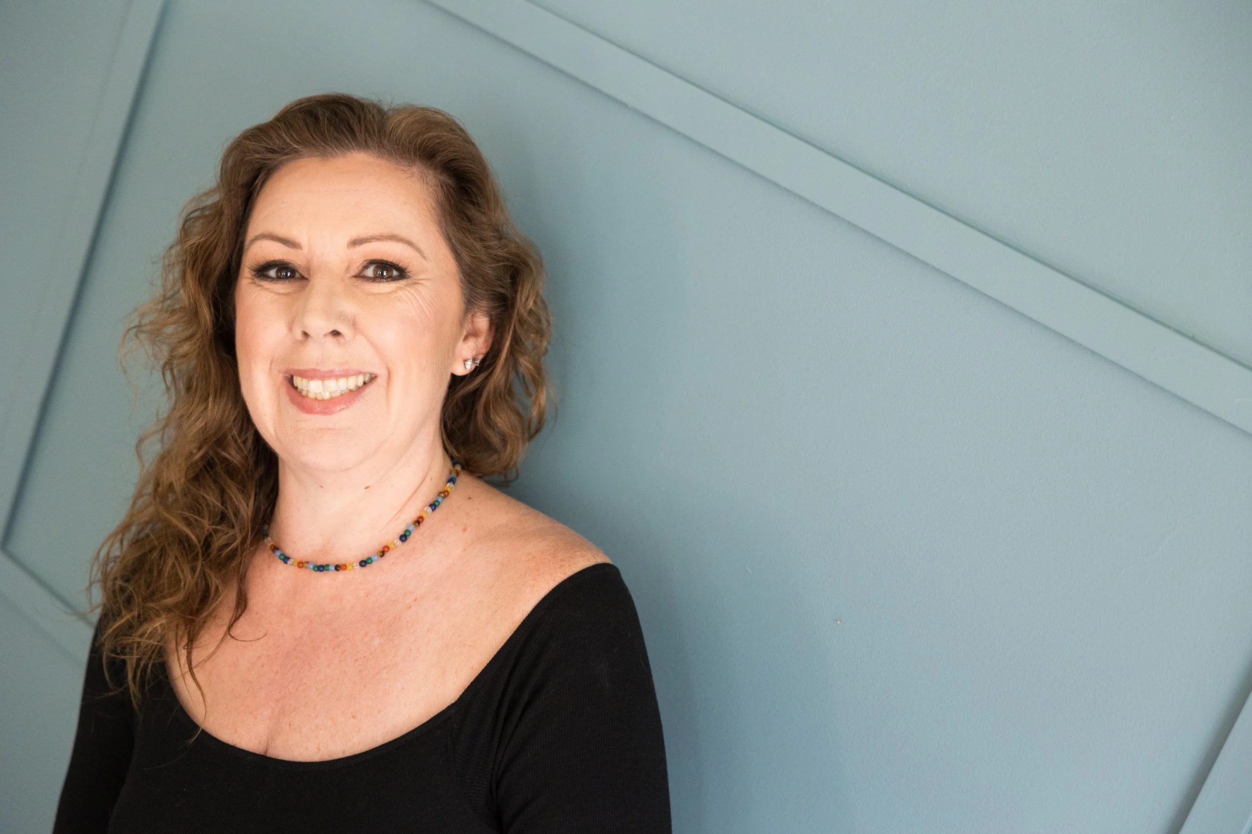 Close-up of a woman with curly, light brown hair, smiling, wearing a black top and a colorful beaded necklace, against a blue wall background.