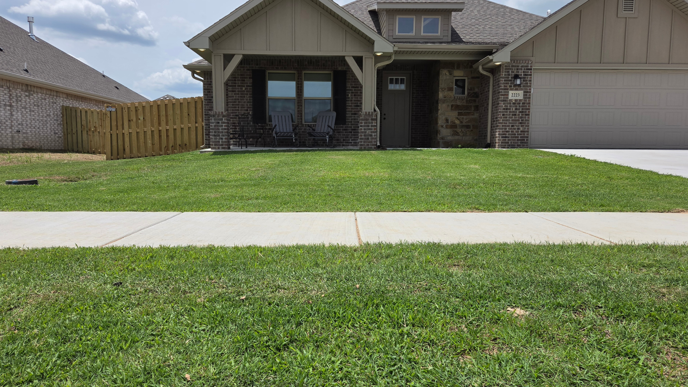 Front view of a suburban house with brick and siding exterior, a concrete driveway, a small porch with two chairs, a fenced yard, and a green lawn under a partly cloudy sky.