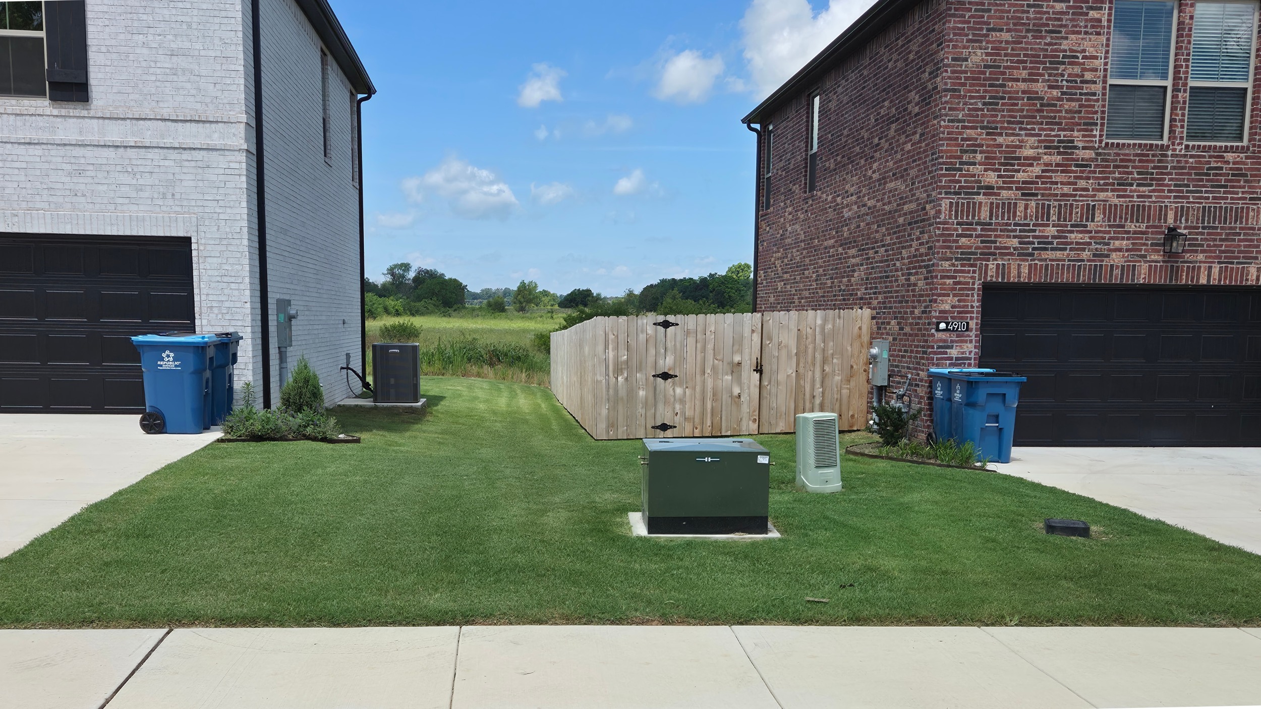 View of a backyard with two houses, a wooden privacy fence, two trash bins, and various utility boxes on the grass and paved surface. Open field and blue sky with clouds in the background.