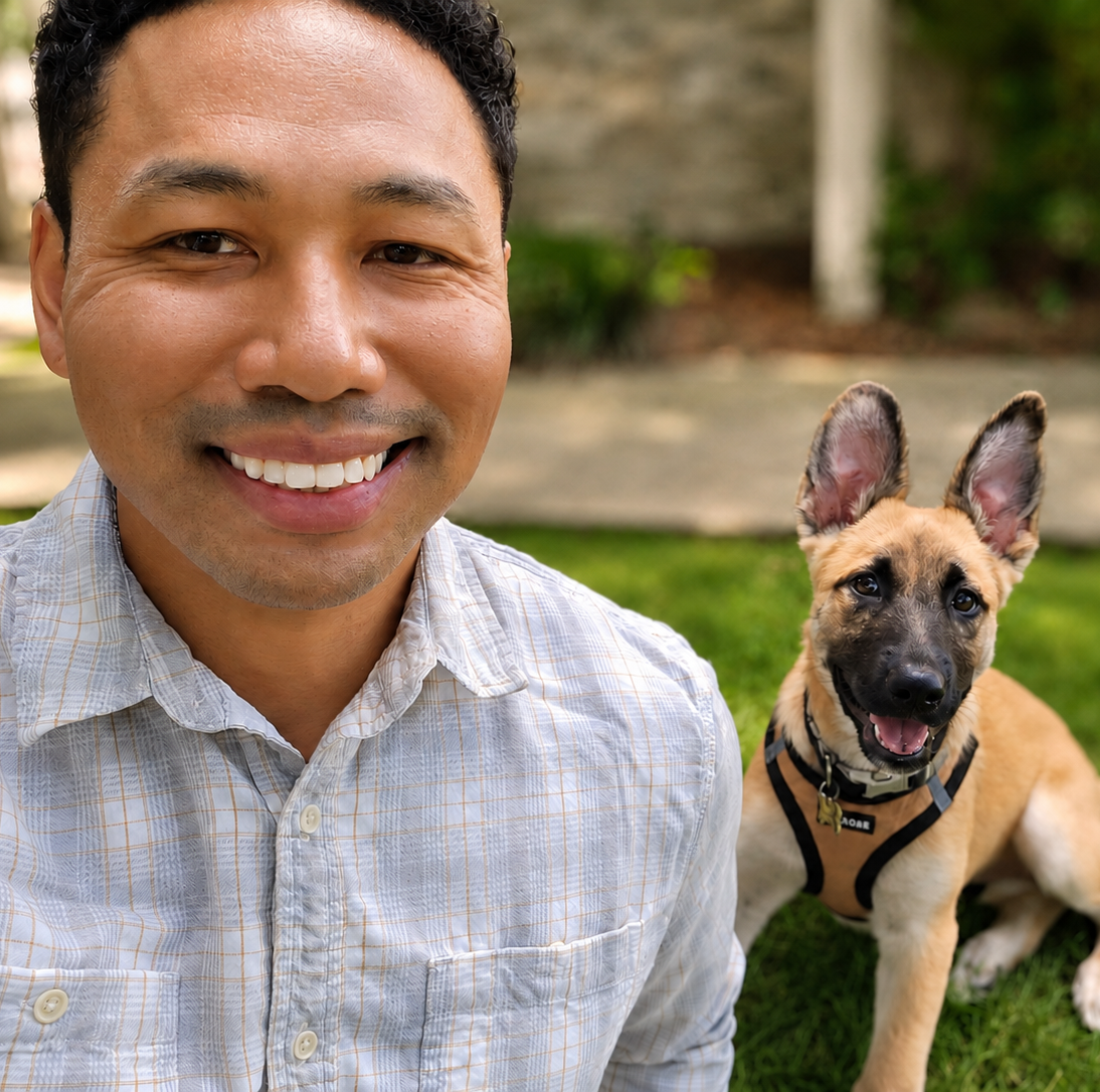 Close-up of a smiling man with a dog sitting on grass in background.