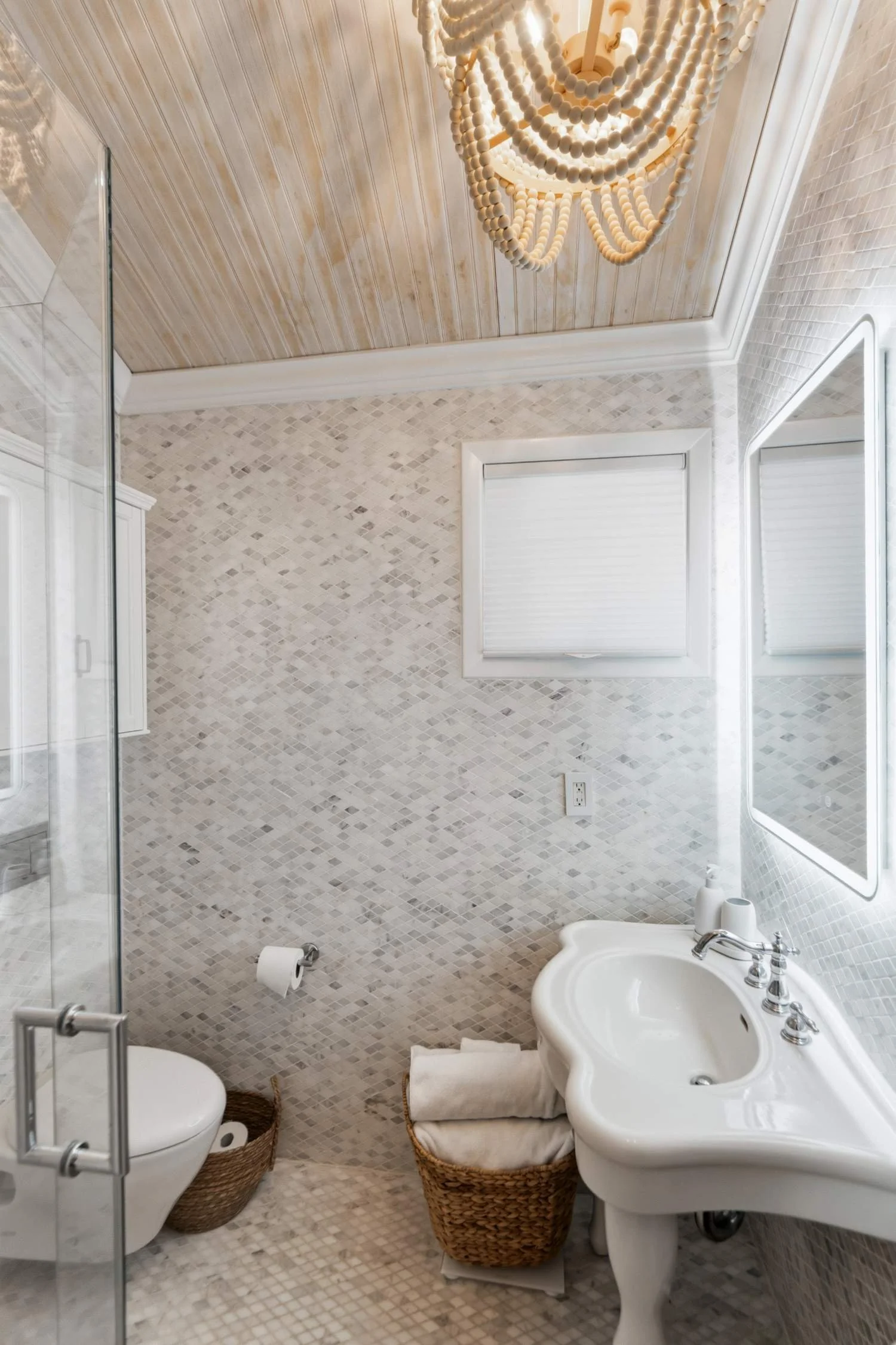A small bathroom with mosaic tile walls and floor, a white porcelain sink with decorative edges, a mirror, a window with blinds, a toilet in a wicker basket, and woven baskets for storage. There is a chandelier hanging from a wooden ceiling.