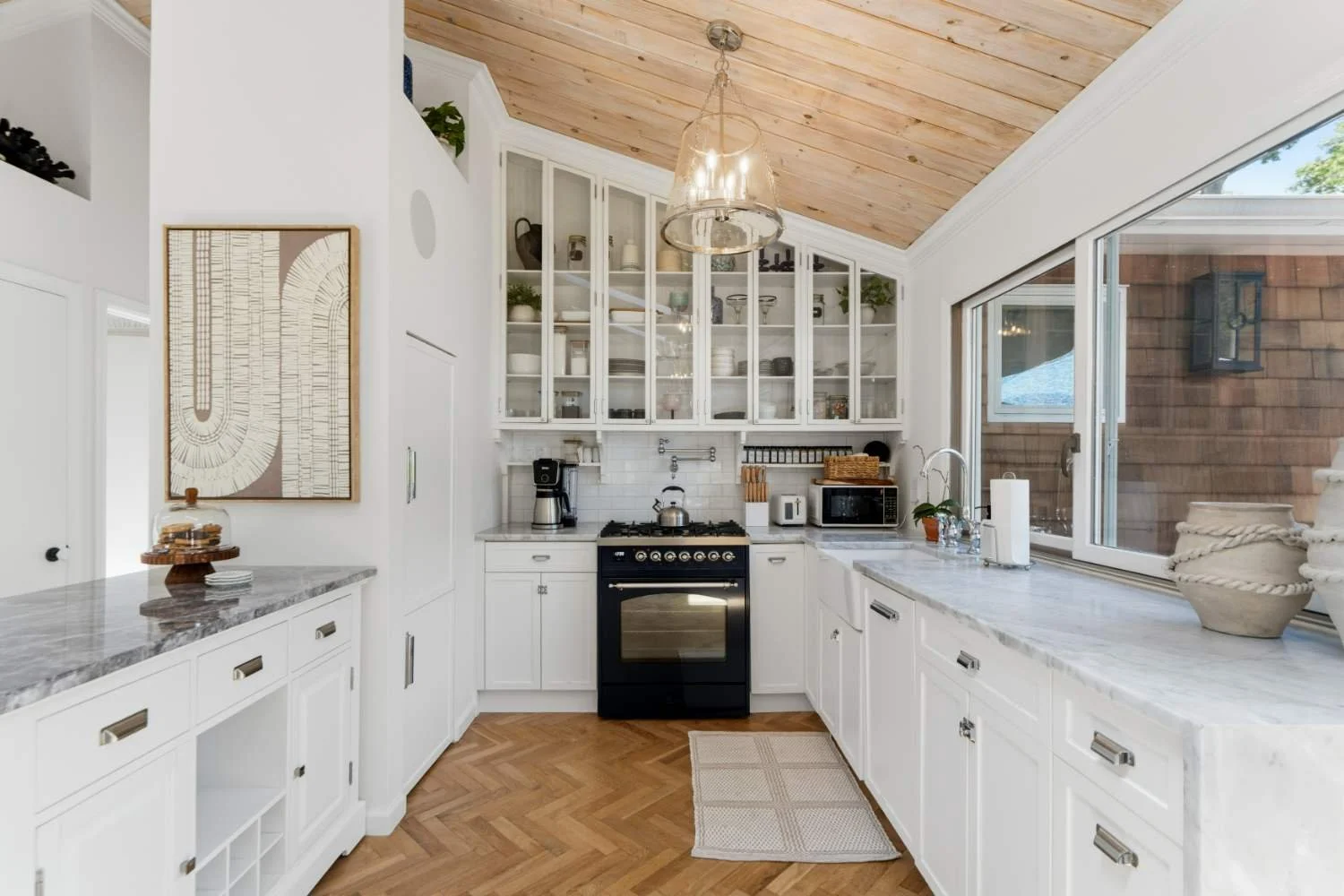 Bright kitchen with white cabinets, granite countertops, and a large window. Wooden ceiling, black stove, coffee maker, microwave, and decorative items on shelves.