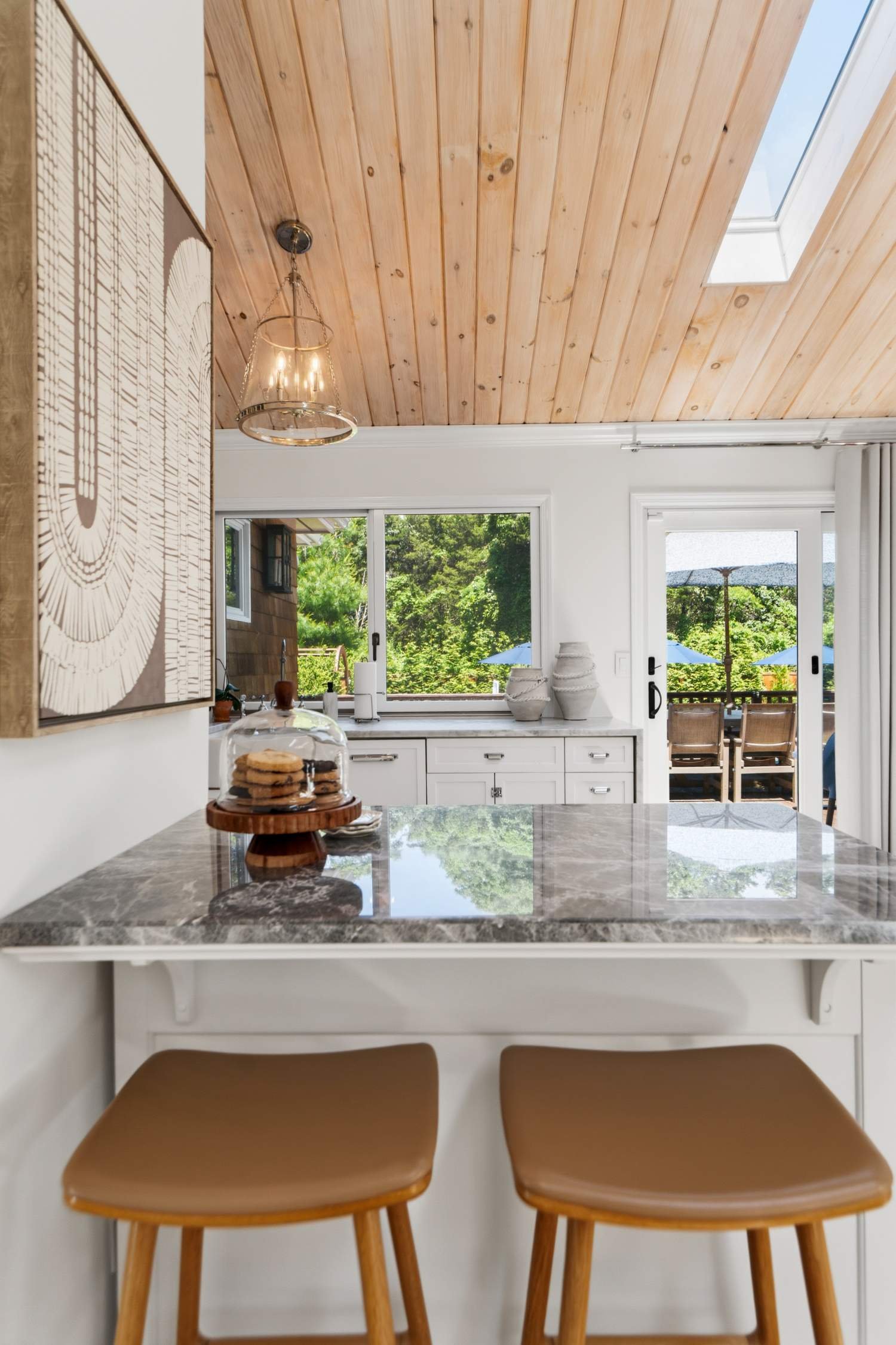 View from inside a kitchen showing a marble countertop with two brown stools underneath, a window with outdoor scenery, and a wooden ceiling with a skylight.