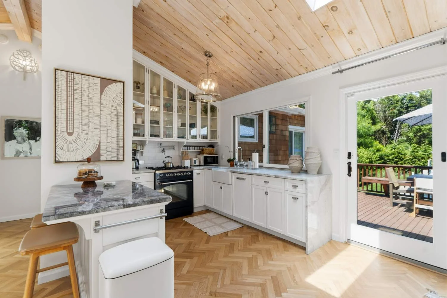 Bright kitchen with white cabinets, a marble countertop, a black stove, and a large sliding glass door opening to a wooden deck with outdoor seating and green trees in the background.