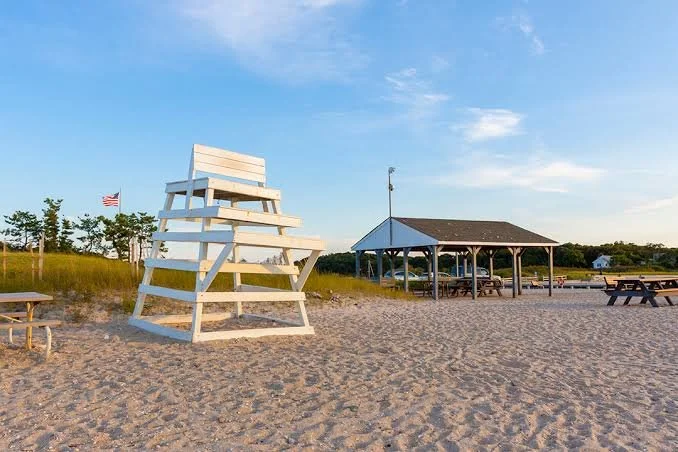 Beach scene with a white lifeguard chair, a pavilion with picnic tables, and an American flag in the distance under a blue sky.
