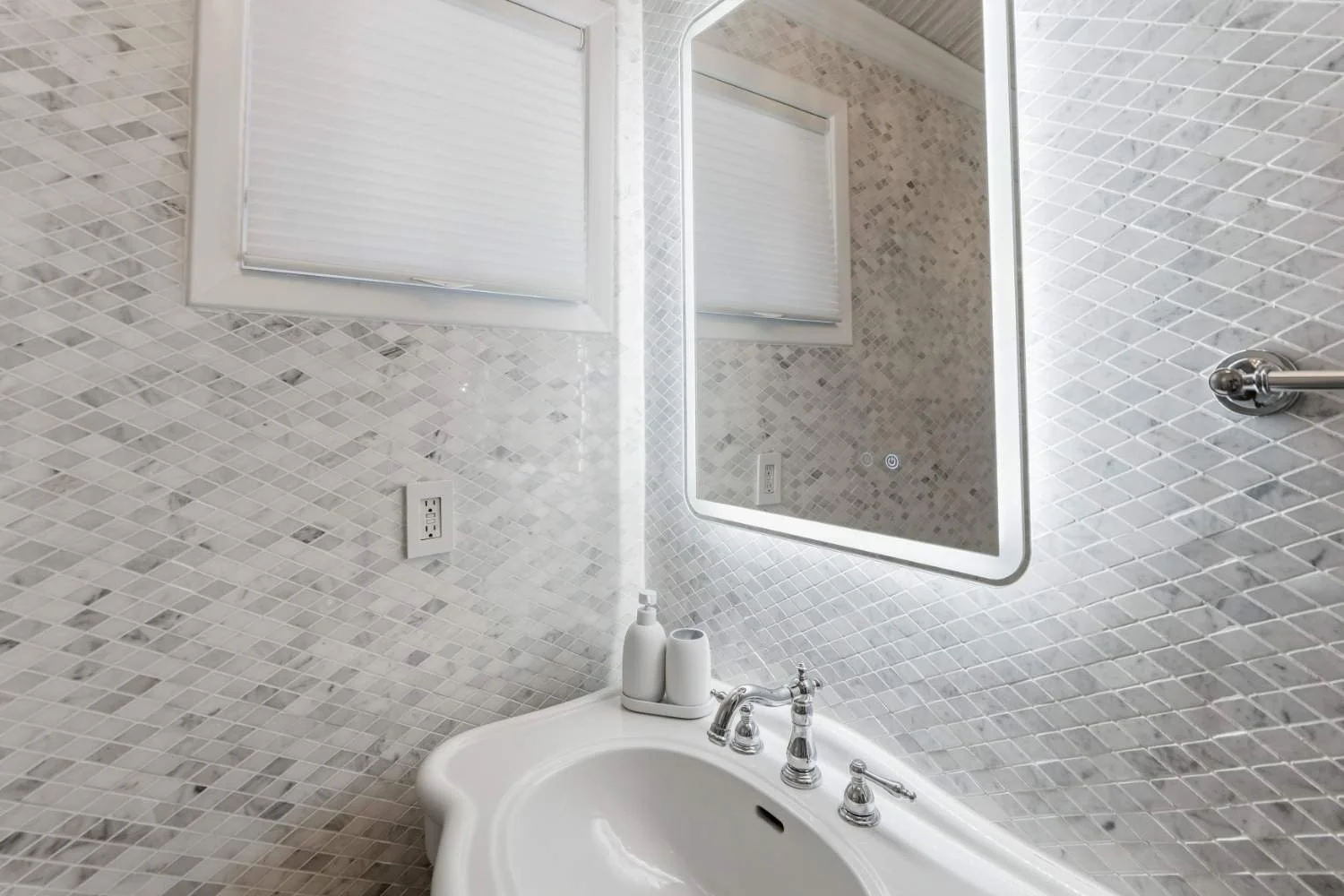 Close-up of a bathroom sink with white soap and lotion dispensers, a mirrored wall with LED lighting, a window with closed blinds, a tiled wall, and a towel bar.