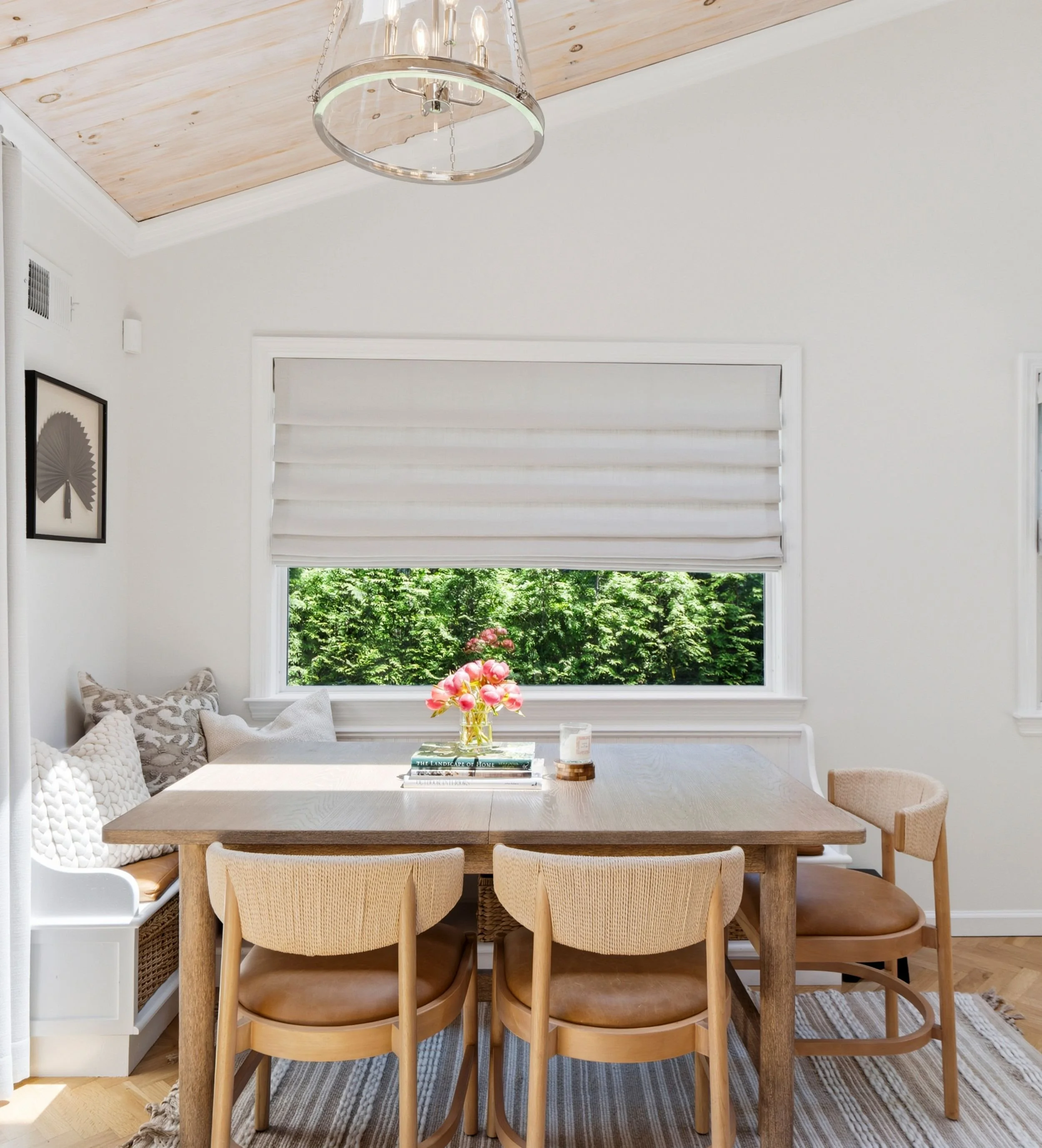 A dining room with a light wooden table surrounded by six beige woven chairs, a large window with white blinds, and a view of green foliage outside. A glass chandelier hangs from the ceiling, and a white bench with patterned cushions is along the left side.
