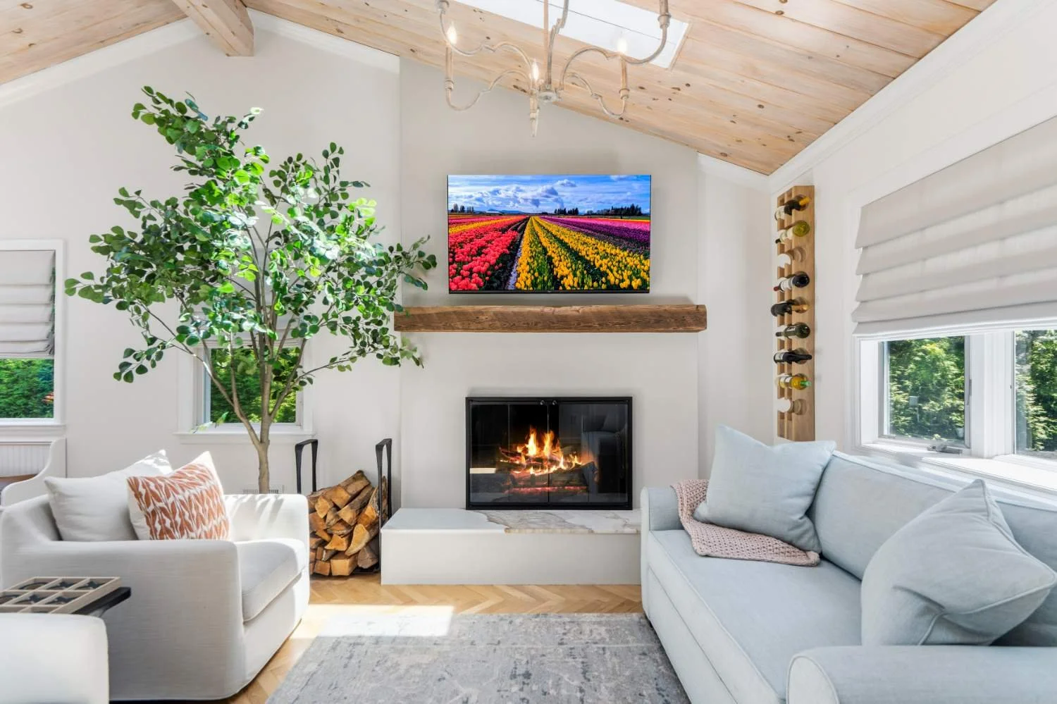 Cozy living room with white couches, a fireplace, a large potted plant, a wall-mounted TV showing colorful tulip fields, a window with blinds, and a wine rack, with a pile of firewood stacked beside the fireplace.