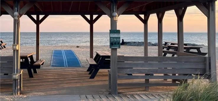 View of a beach pavilion with wooden picnic tables, a walkway leading to the beach, and the ocean in the background at sunset.
