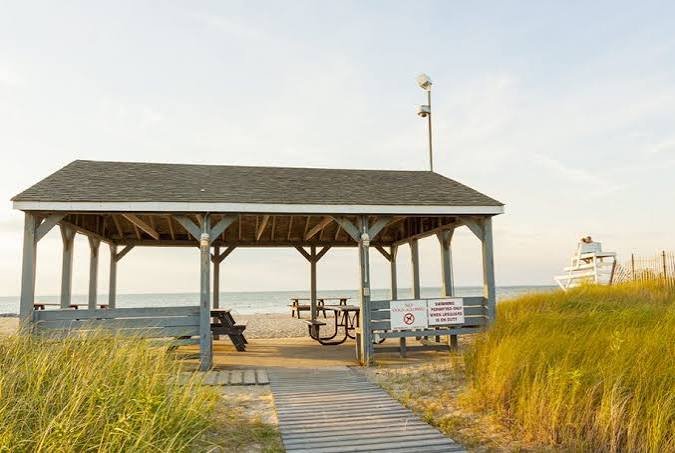 A seaside pavilion with picnic tables and a wooden walkway leading to the beach, surrounded by grassy dunes and a lifeguard tower in the background.