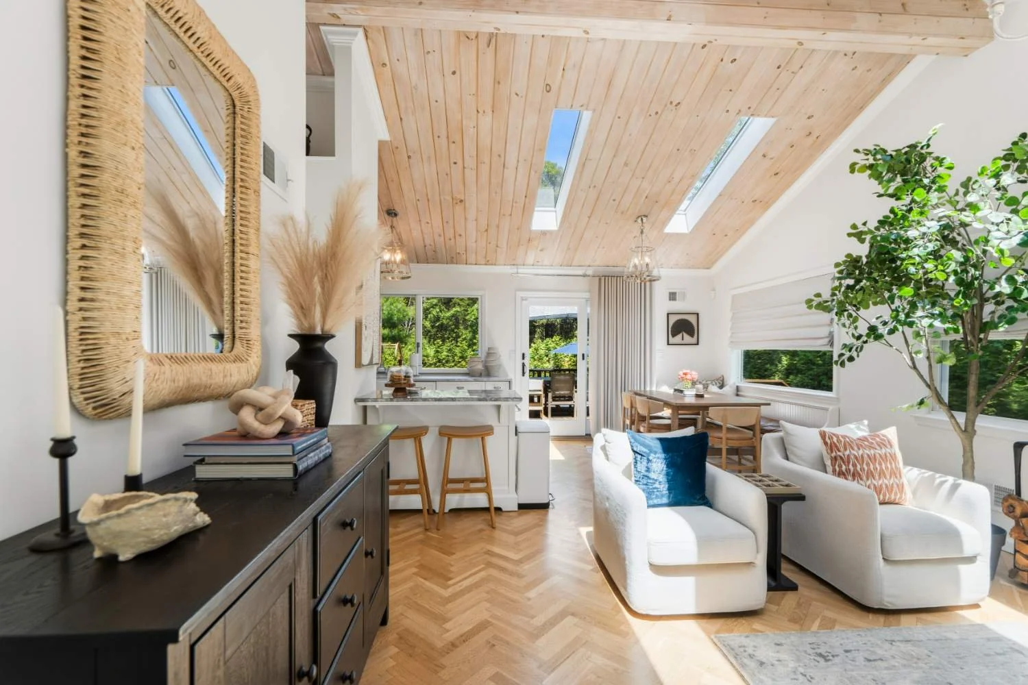 Bright living room with white sofa, blue and orange pillows, wooden dining table, large windows, skylights, wooden ceiling, and green indoor plant.