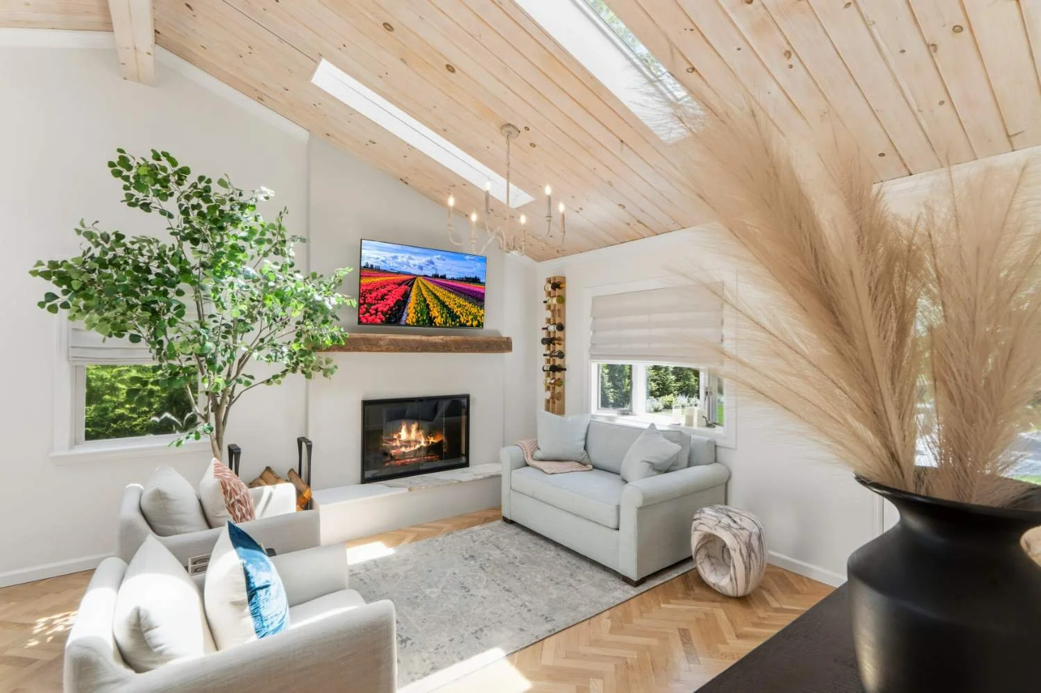 Bright living room with white walls, wooden ceiling and floor, a fireplace, a large window, plants, and a television displaying colorful fields.