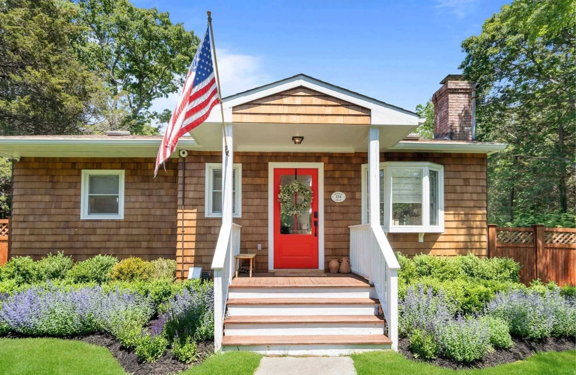 Front view of a house with a red front door, white porch steps, a porch light, a wreath on the door, an American flag on a pole, two clay pots, small shrubs, and purple flowers in the garden, surrounded by green trees.