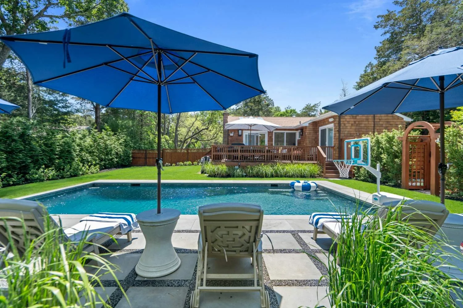 Backyard with a swimming pool, surrounded by lounge chairs and umbrellas, with a wooden deck and house in the background.